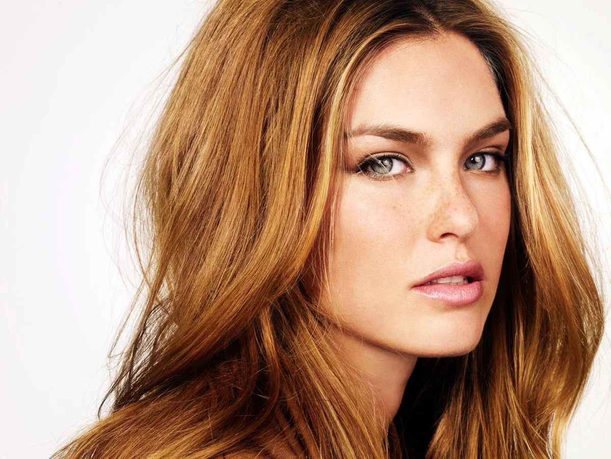 Close-up of a woman with long, wavy red hair and blue eyes against a white background.