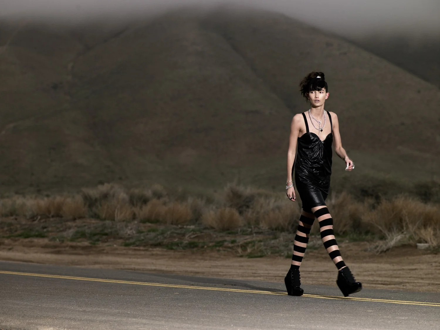 A woman walking on a deserted road in a mountainous area, wearing a black dress, striped stockings, and platform shoes.