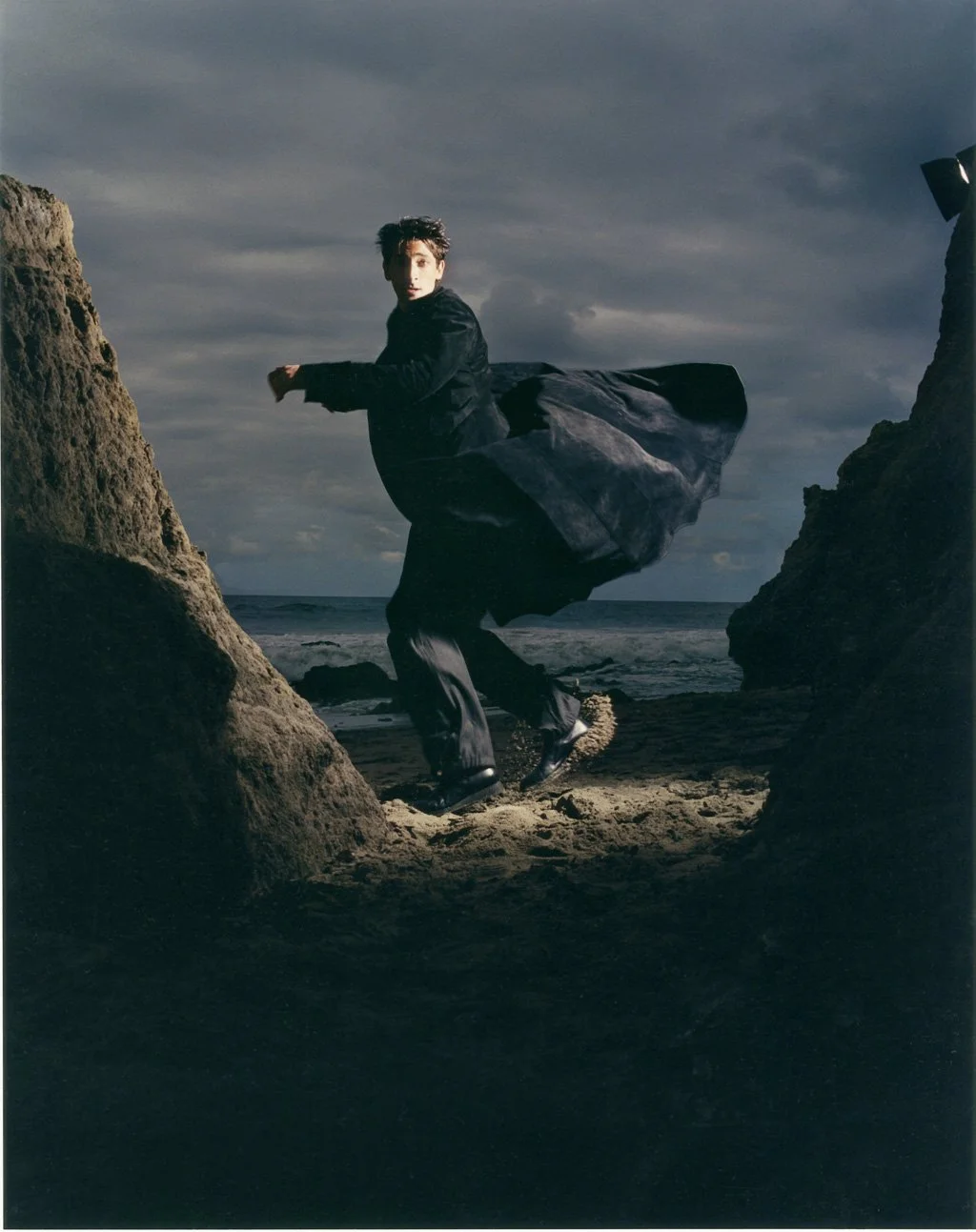 ADRIEN BRODY wearing a black coat and pants, running along a beach between two large rocks with the ocean in the background under cloudy sky.