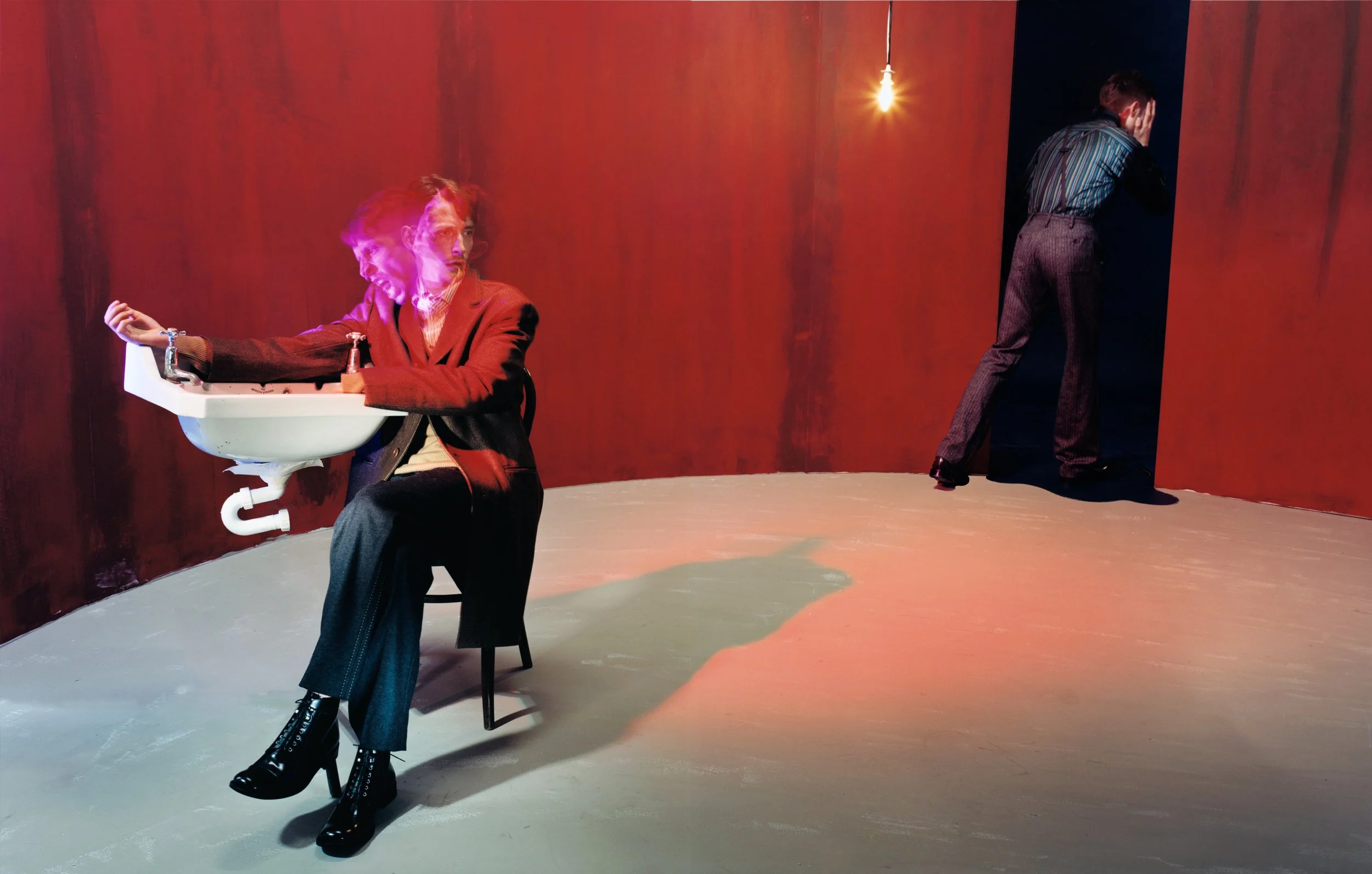 Two individuals in a room with red walls and a concrete floor. One person is sitting on a chair, holding a white sink in their lap, and appears to be glitching or experiencing digital distortion. The other person is standing in the corner with their 