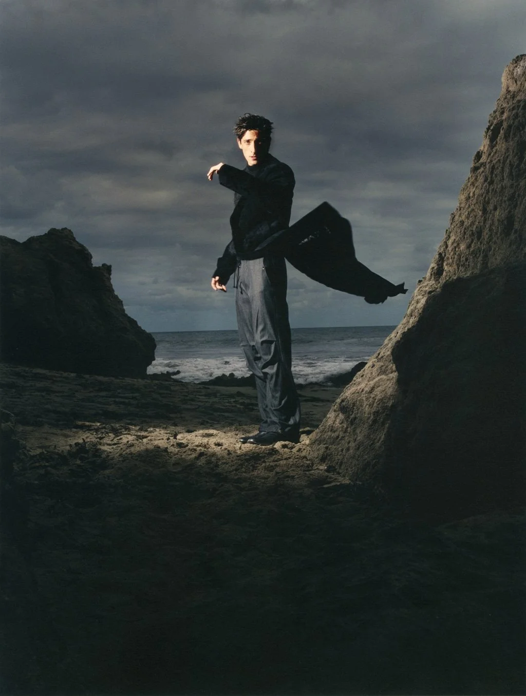 ADRIEN BRODY standing on a beach between rocks with a stormy sky in the background, wearing dark clothing and looking at the camera.