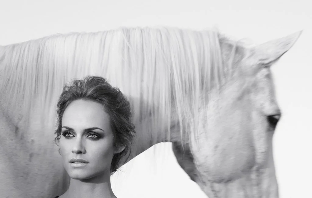 A woman with styled hair and makeup, and a white horse with a flowing mane, both in a black and white portrait.