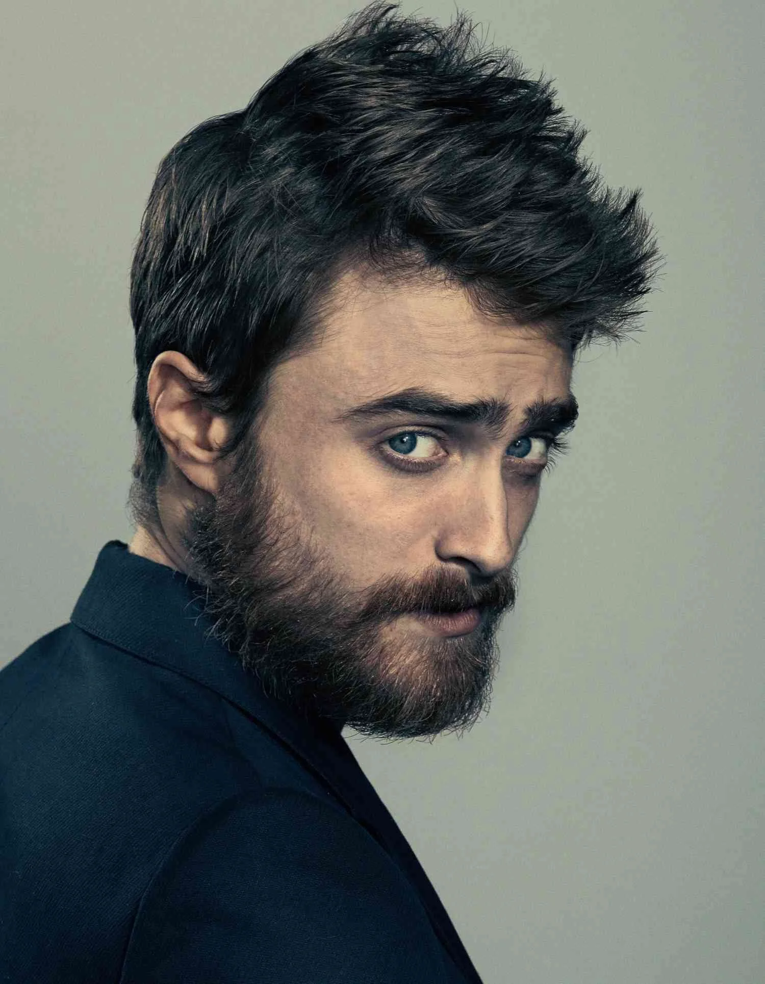 Close-up of DANIEL RADCLIFFE with dark hair and beard, looking over his shoulder at the camera against a plain background.