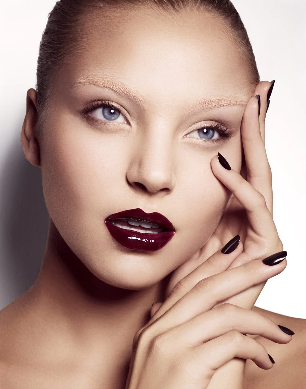 Close-up of a woman's face with striking makeup, dark lipstick, light eyes, and manicured nails, touching her face with her fingers.