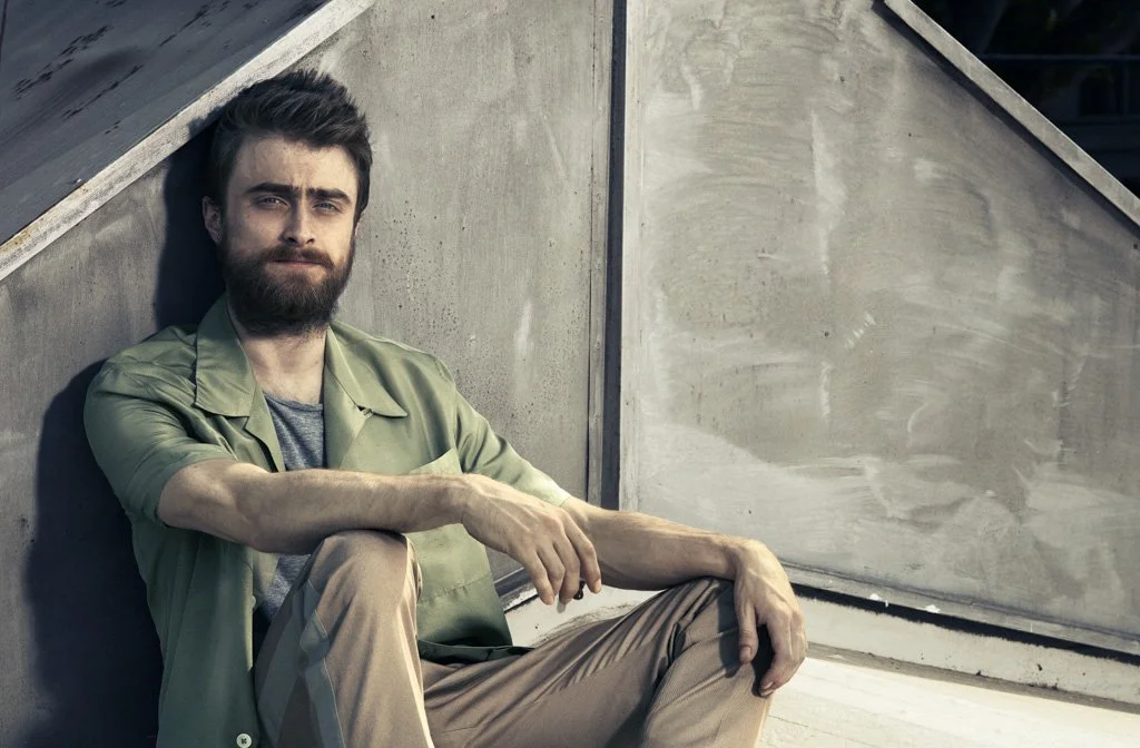 DANIEL RADCLIFFEwith brown hair, beard, and wearing a green shirt over a gray T-shirt, sitting on concrete steps, leaning against a wall.