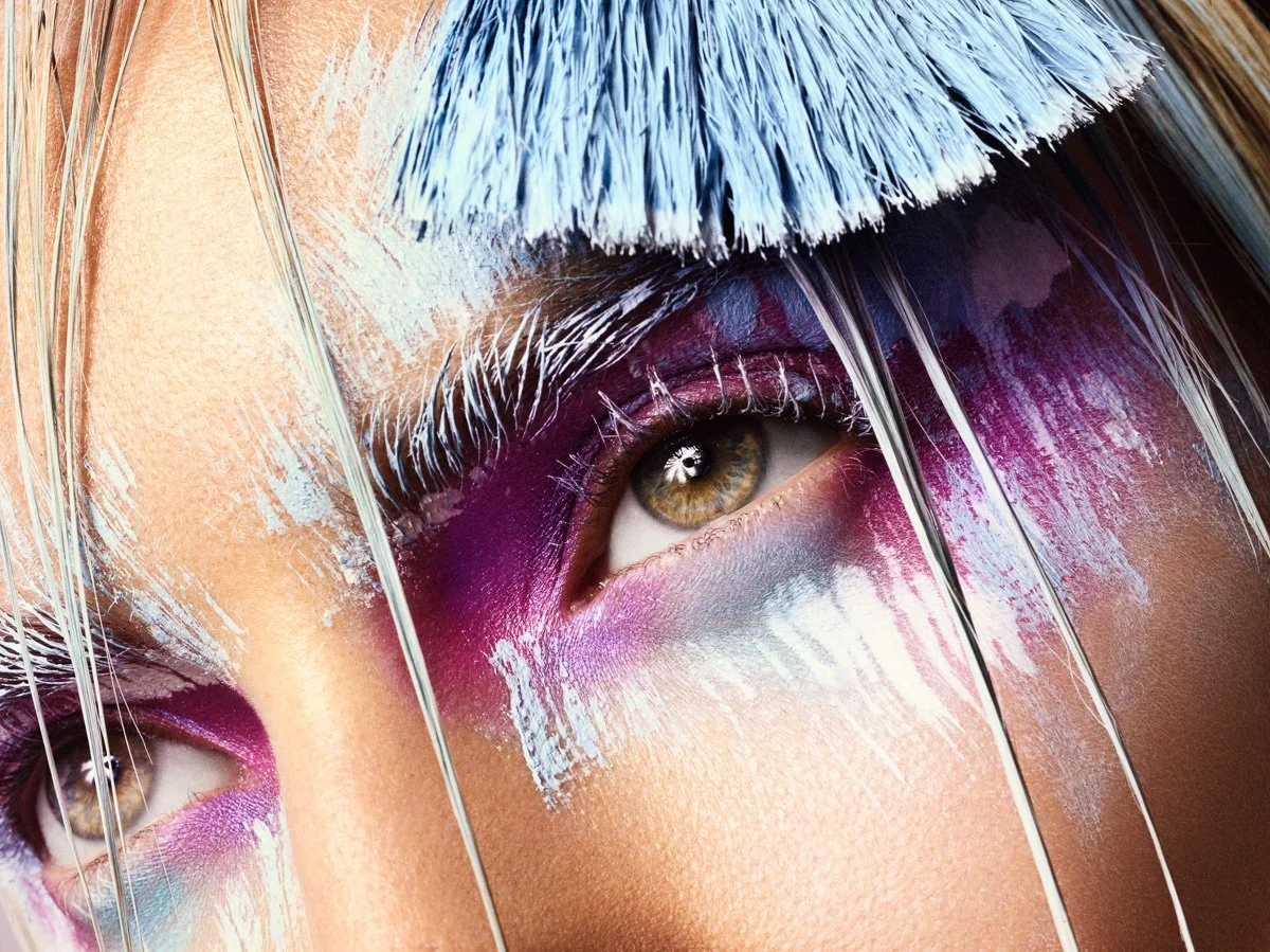 Close-up of a person's face with artistic makeup, including purple, pink, and blue eyeshadow, and white and silver streaks on their eyebrows and eyelids.