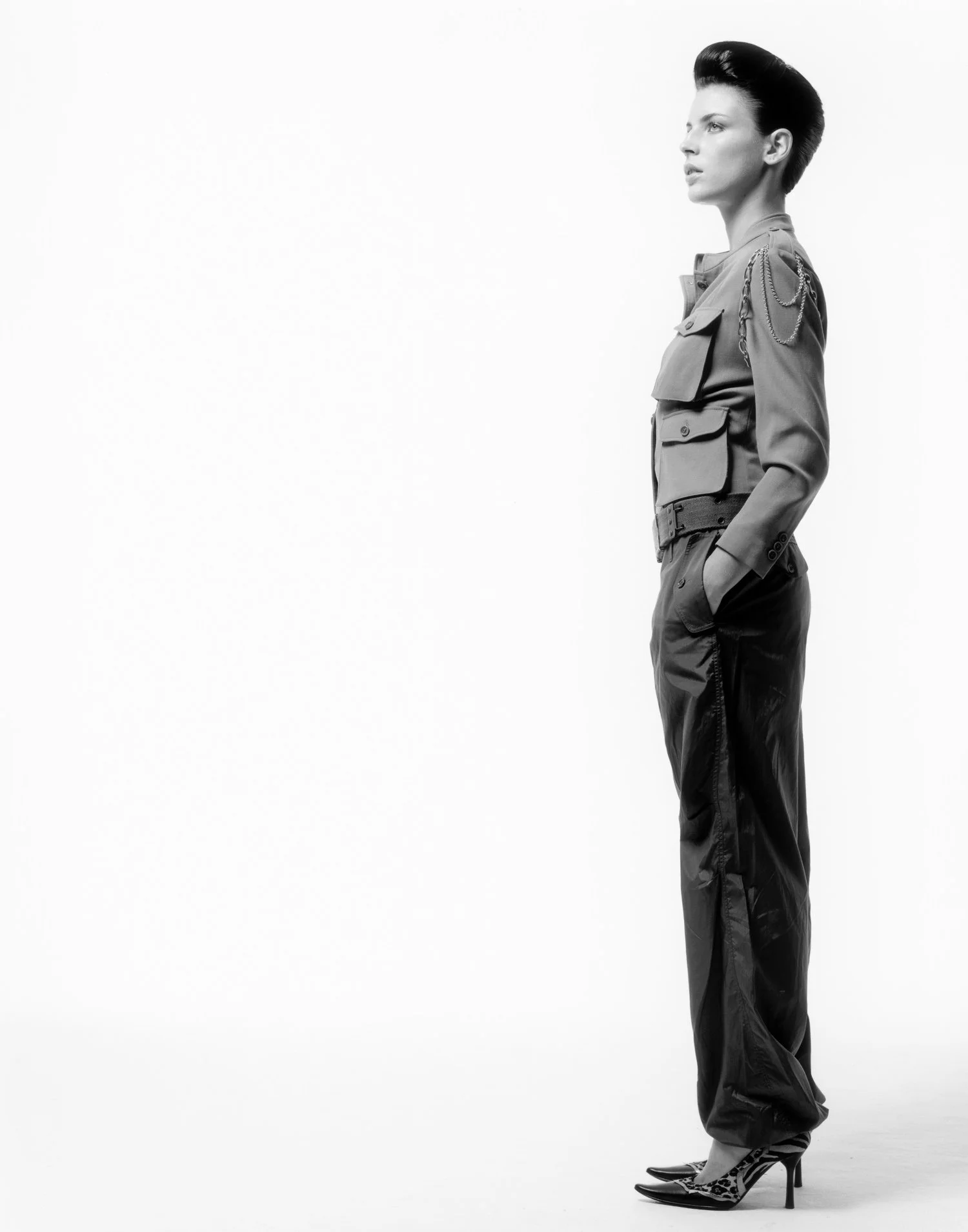A woman standing in profile, dressed in a stylish military-inspired outfit with high heels, against a plain white background.