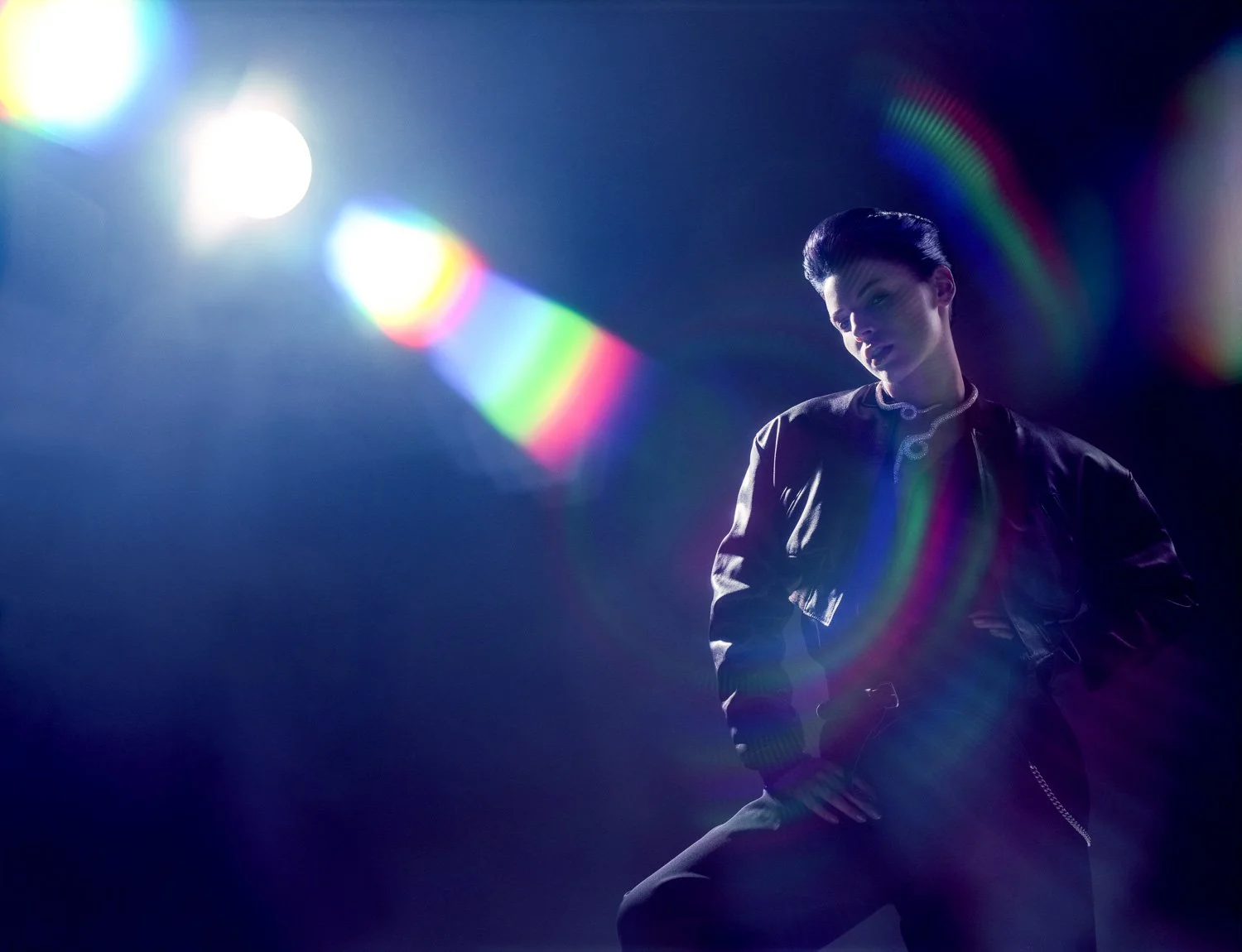 A person with dark, slicked-back hair wearing a black leather jacket, sitting against a dark background with bright lights creating rainbow lens flares.