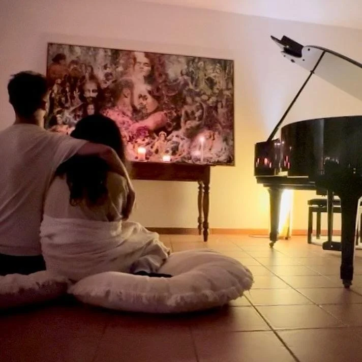 A couple sits on floor cushions facing a painting, with a grand piano nearby, in a cozy room.