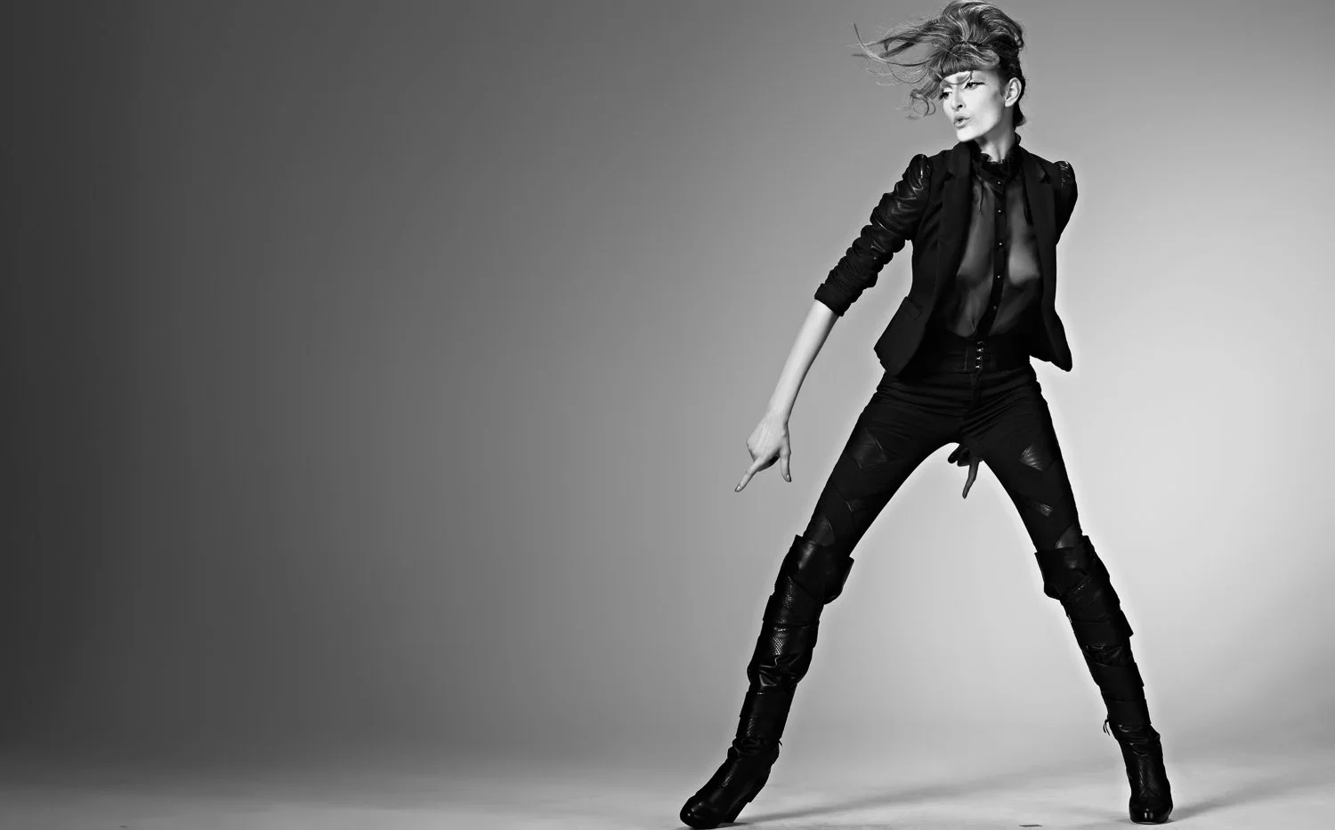 Black and white photo of a woman in stylish clothing, standing with legs apart and pointing downward with her right hand. Her hair is styled with a lot of volume and blows to the left.