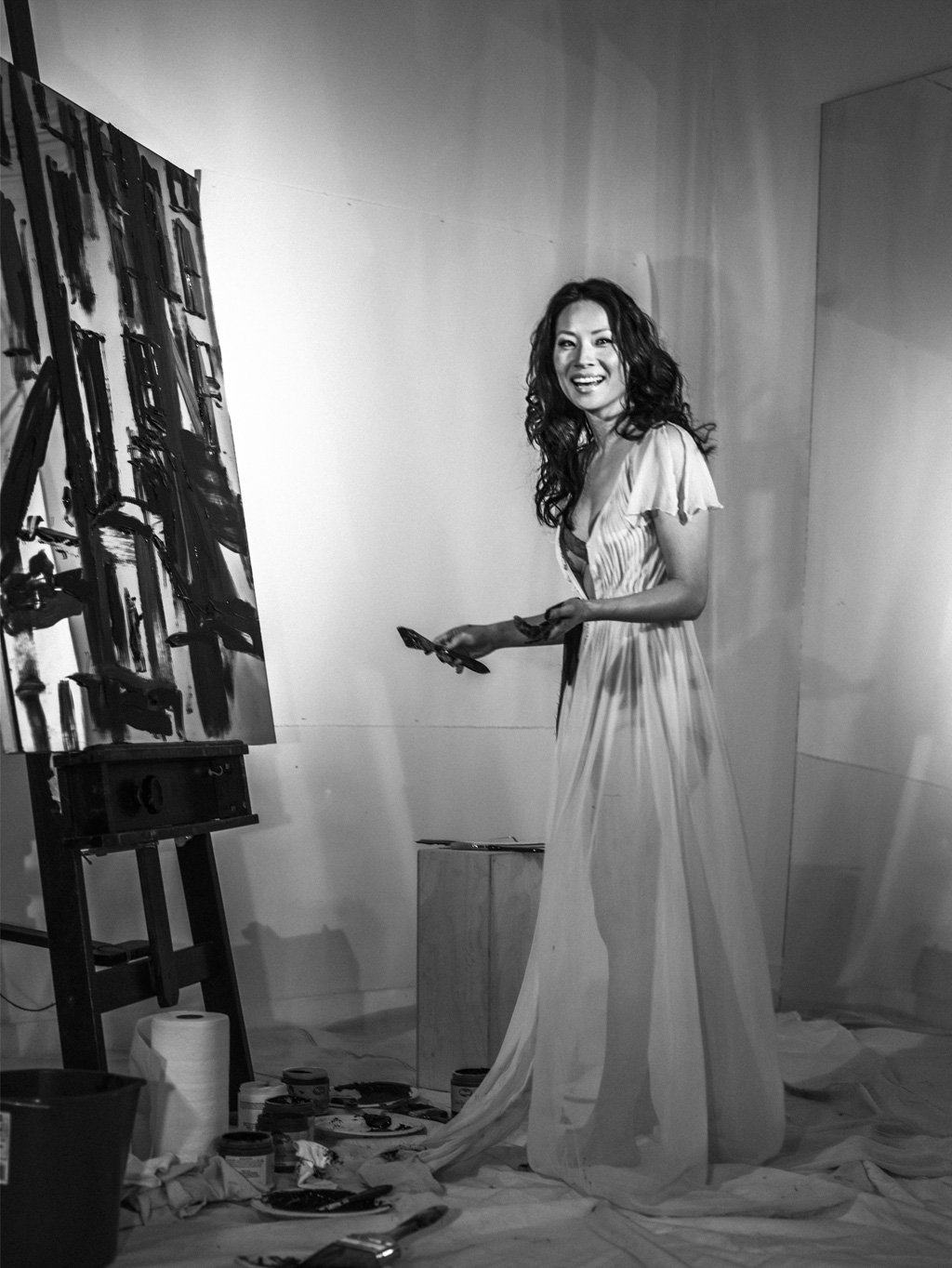 LUCY LIU painting a large abstract black and white artwork on a canvas, standing in an art studio with painting supplies and a roll of paper towels on the floor.