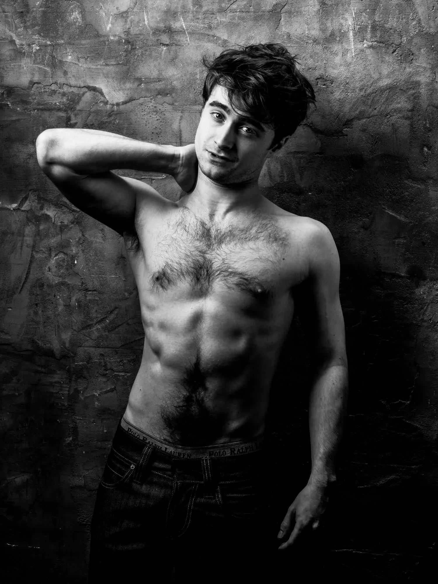 A shirtless DANIEL RADCLIFFEwith messy hair, standing against a textured wall, looking into the camera with a slight smile, one hand behind his neck.