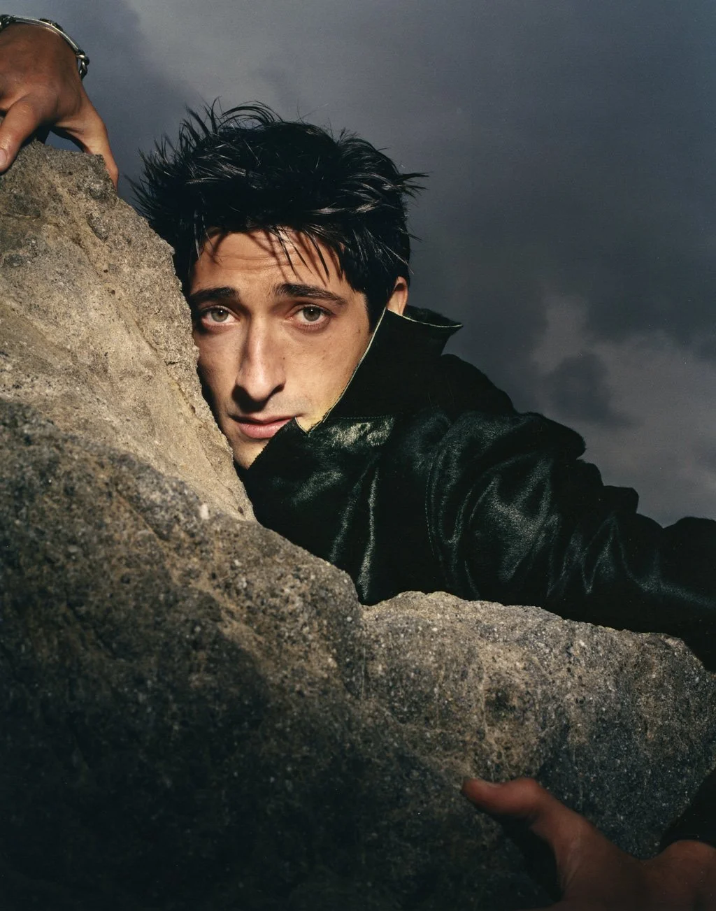 ADRIEN BRODY with messy black hair and a black leather jacket is peering over a large rock, with dark stormy clouds in the background.