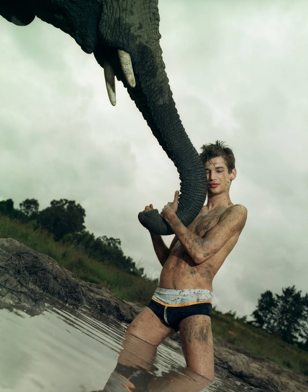 A shirtless young man with messy hair and mud on his body holding a large African elephant's trunk near a river with a cloudy sky in the background.