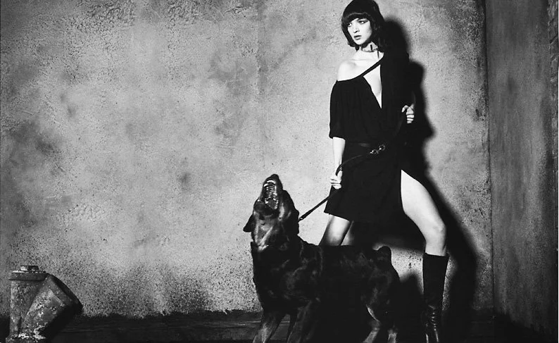 A woman with short hair wearing an off-the-shoulder black dress, standing against a textured wall, holding a leash attached to a large dog.