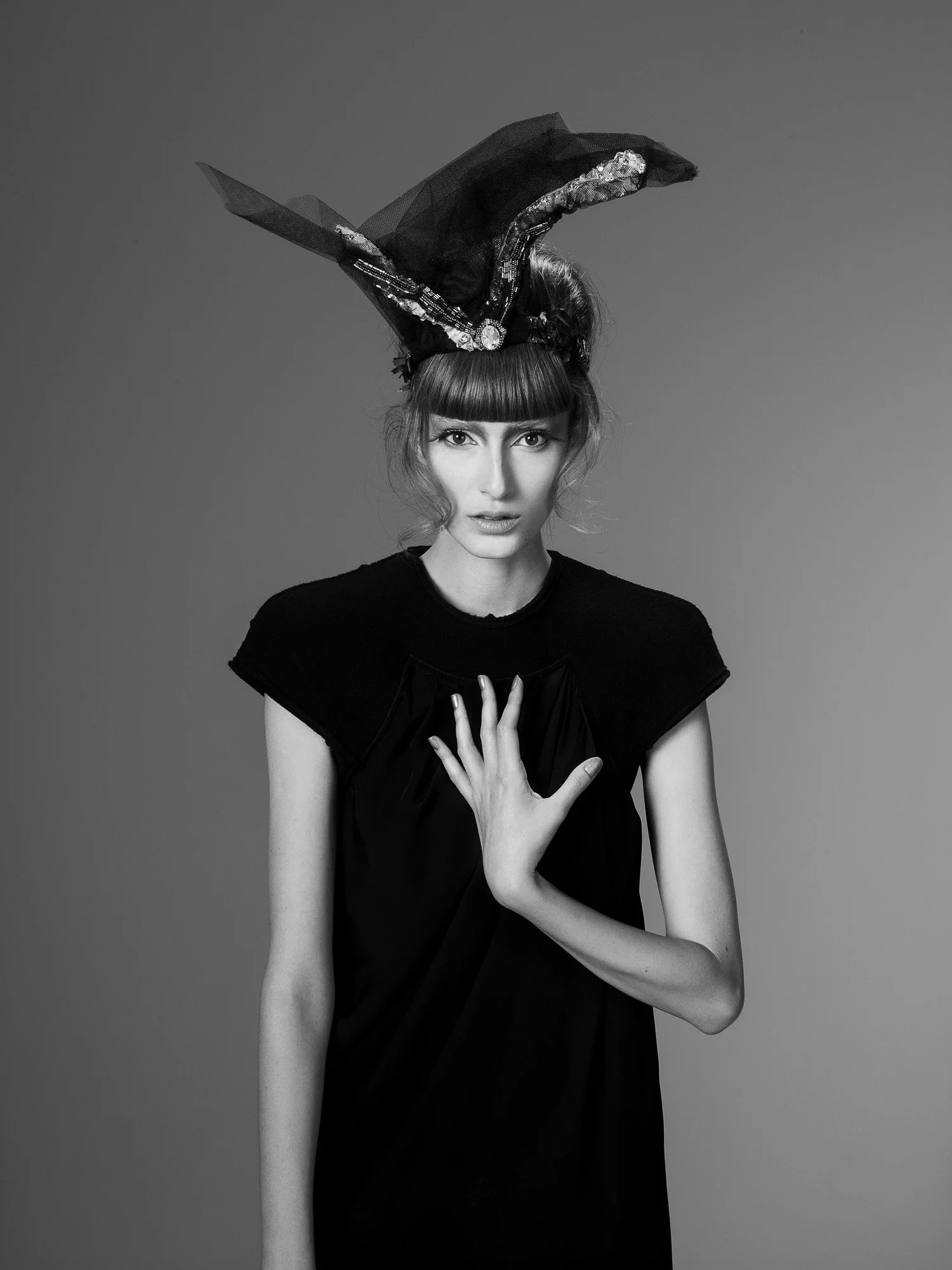 A woman wearing a large, elaborate black headpiece with sequins and tulle, and a black dress or top, holding one hand to her chest against a plain background.