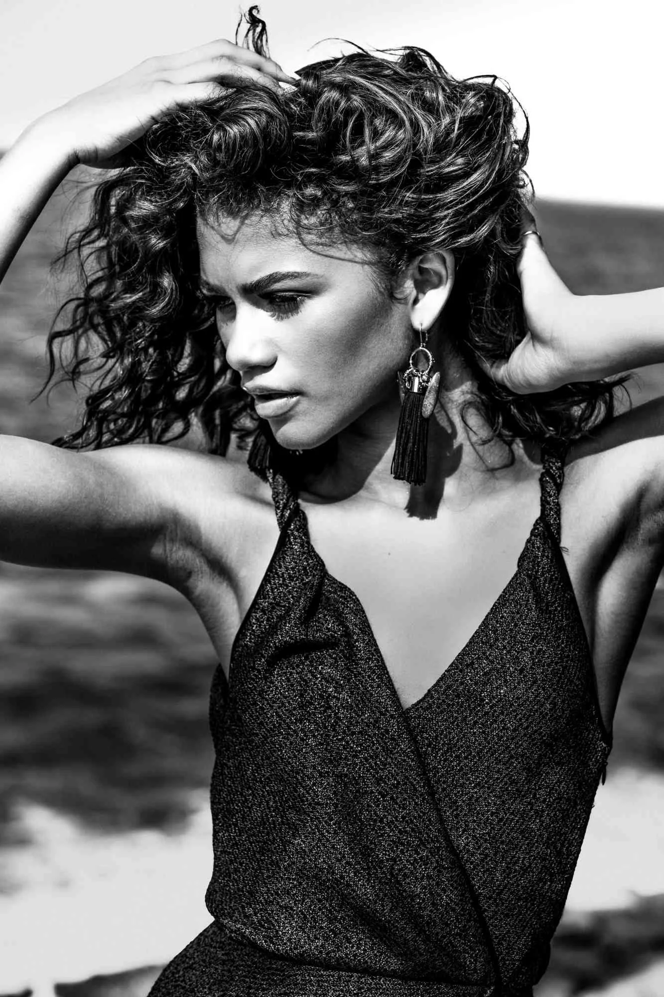 ZENDAYA with curly hair posing outdoors, wearing a sleeveless top and statement earrings, with a focused expression.