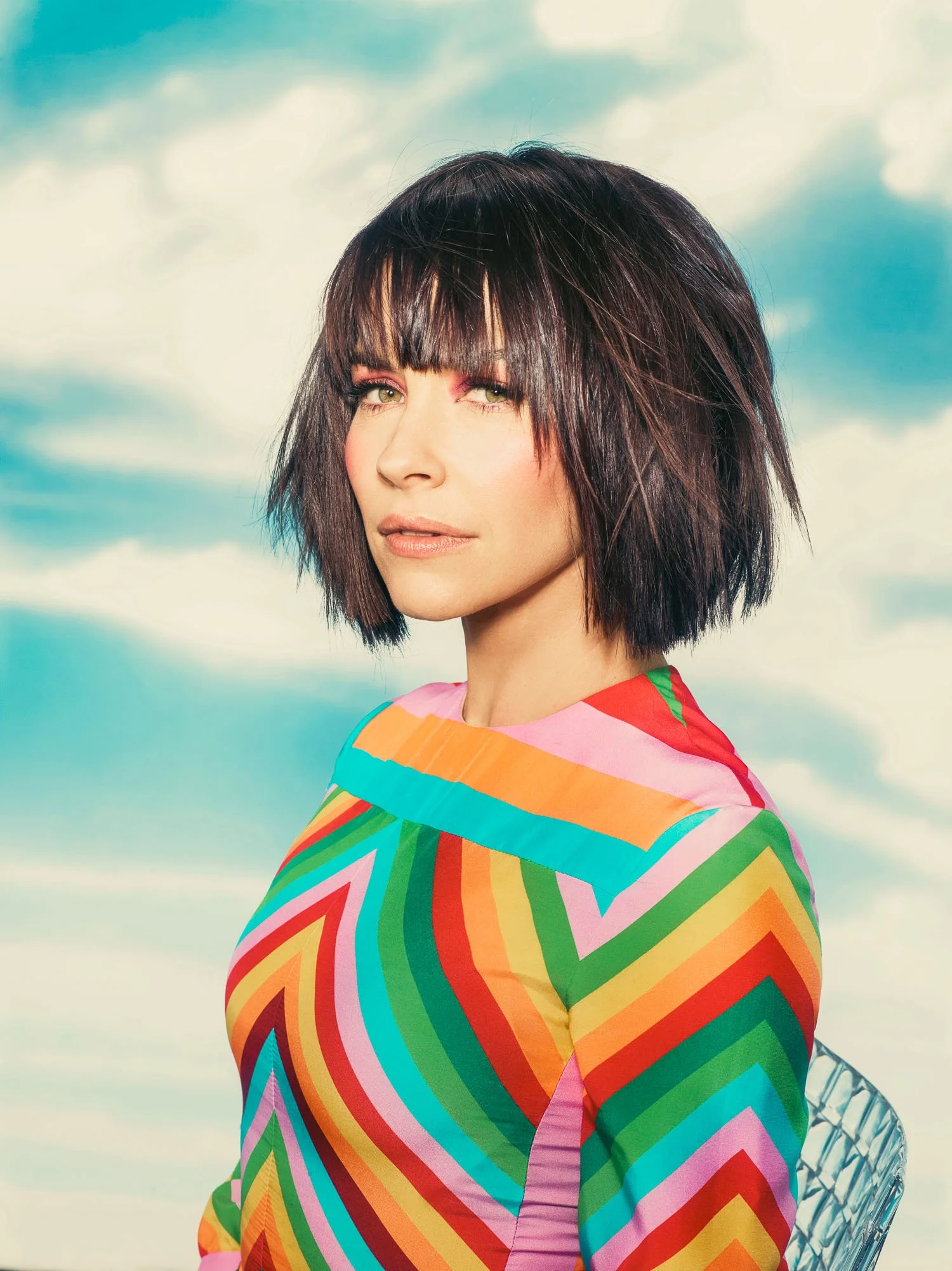 EVANGELINE LILLY with short dark hair and bangs, wearing a colorful, zigzag-patterned dress, stands outdoors against a backdrop of a cloudy sky.