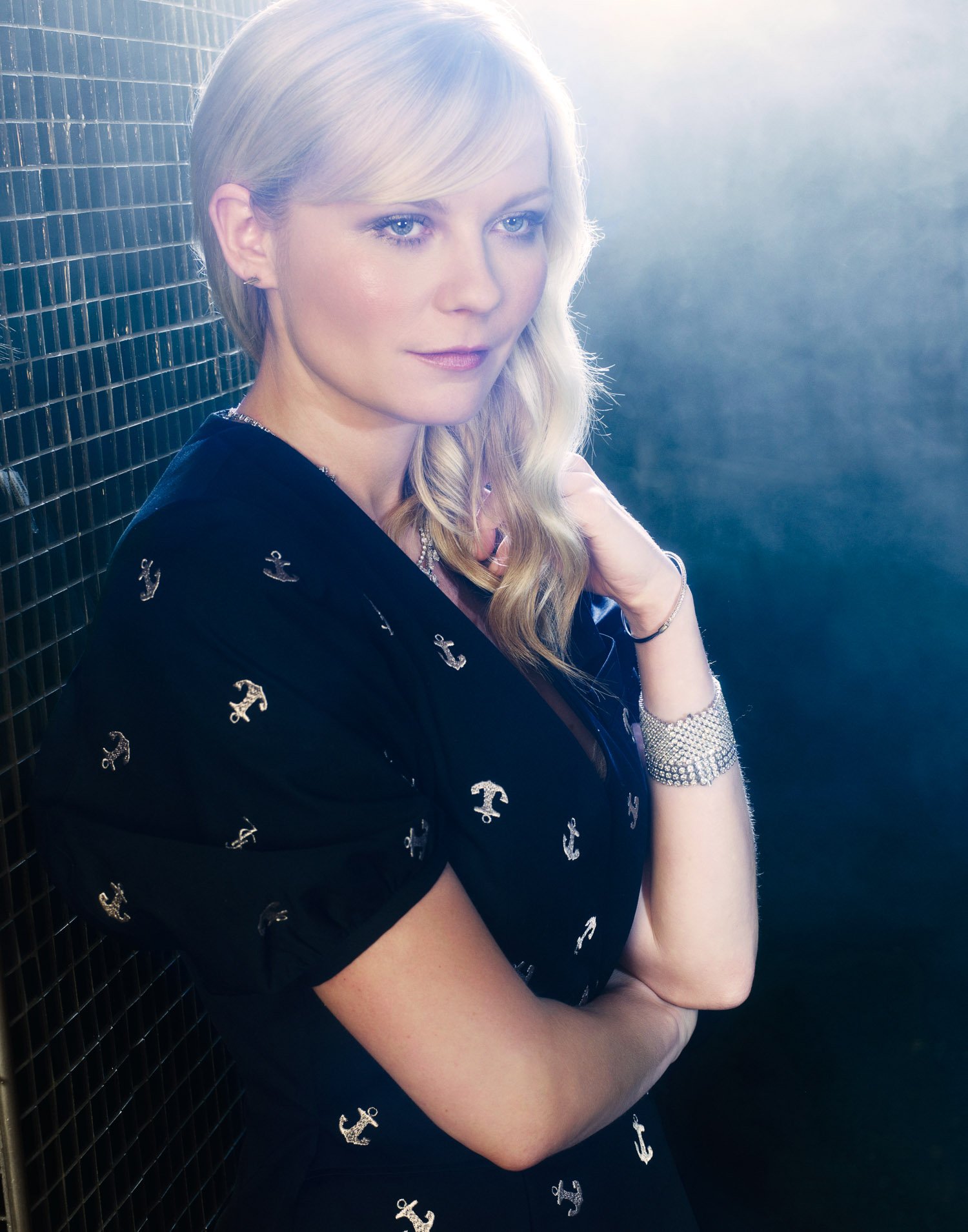 KIRSTEN DUNST with wavy hair posing against a dark tiled wall, illuminated by bright light from behind, wearing a black dress with anchor patterns and a sparkly bracelet.