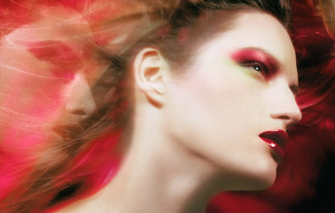 A woman with vibrant makeup, including red eyeshadow, green eyeshadow, and glossy red lipstick, lying with her head turned sideways surrounded by flowing red and pink hair.