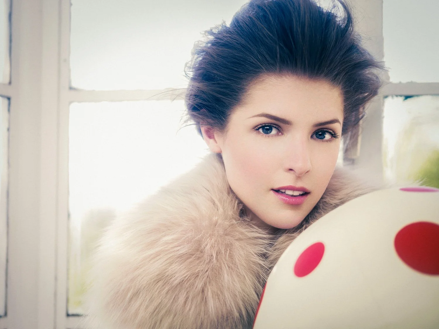 ANNA KENDRICK with short dark hair, blue eyes, and fair skin looking at a white cake with red polka dots, near a window with white trim.