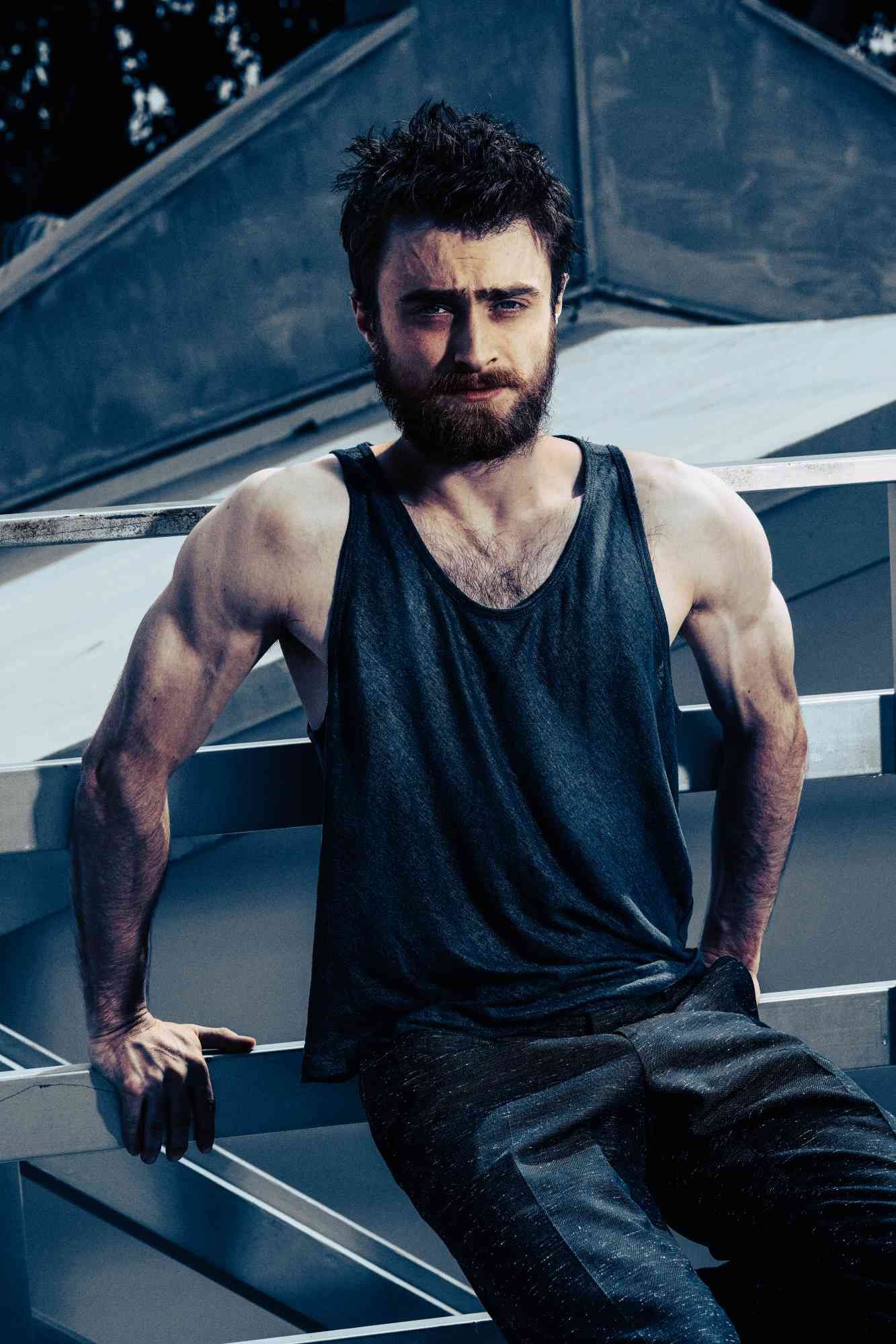DANIEL RADCLIFFE with a beard and messy hair, dressed in a wet black tank top and dark pants, sitting on a metal railing outdoors during daytime.