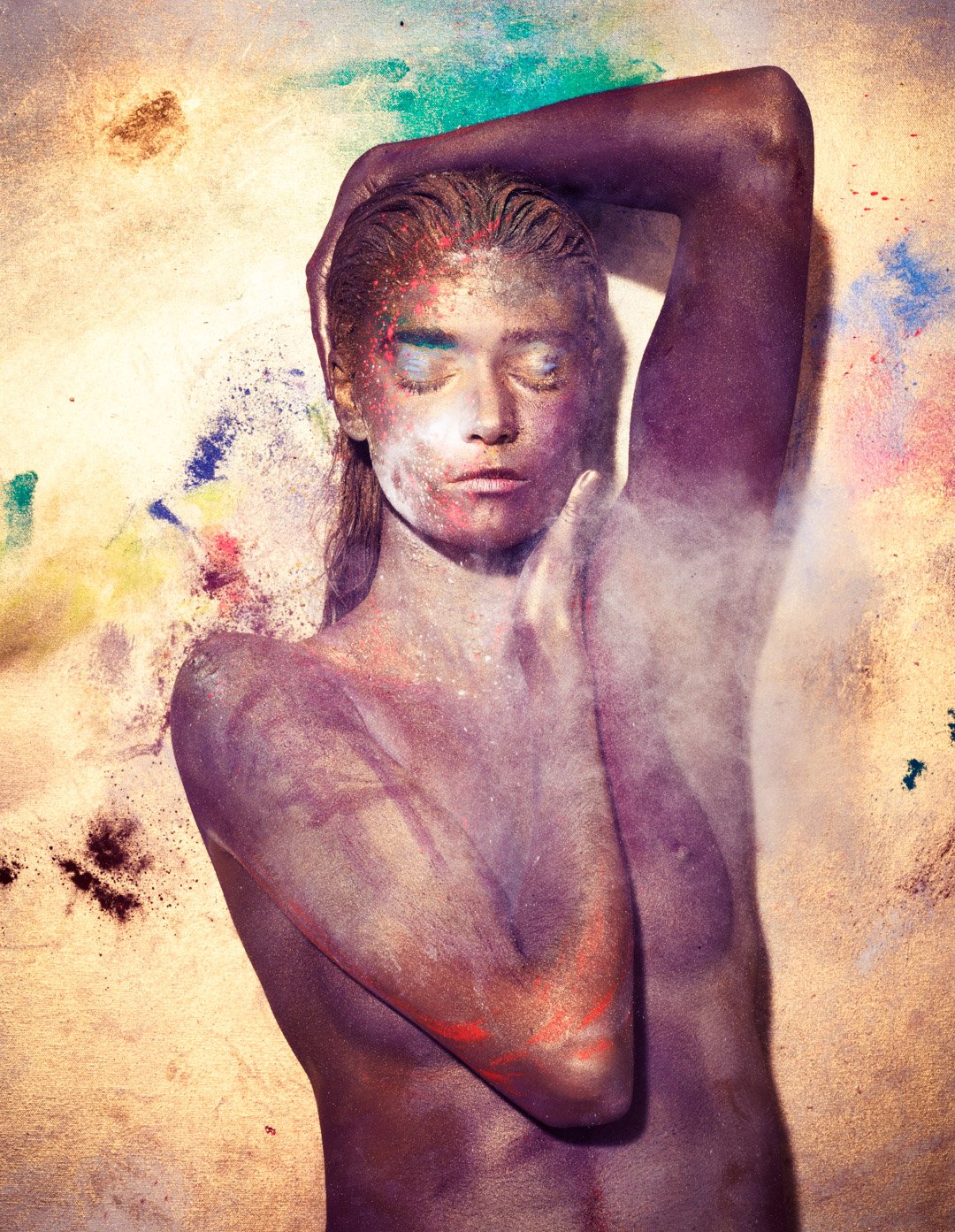 A woman with her eyes closed, covered in colorful powder with her hand resting on her head, surrounded by a vibrant splatter of colors.