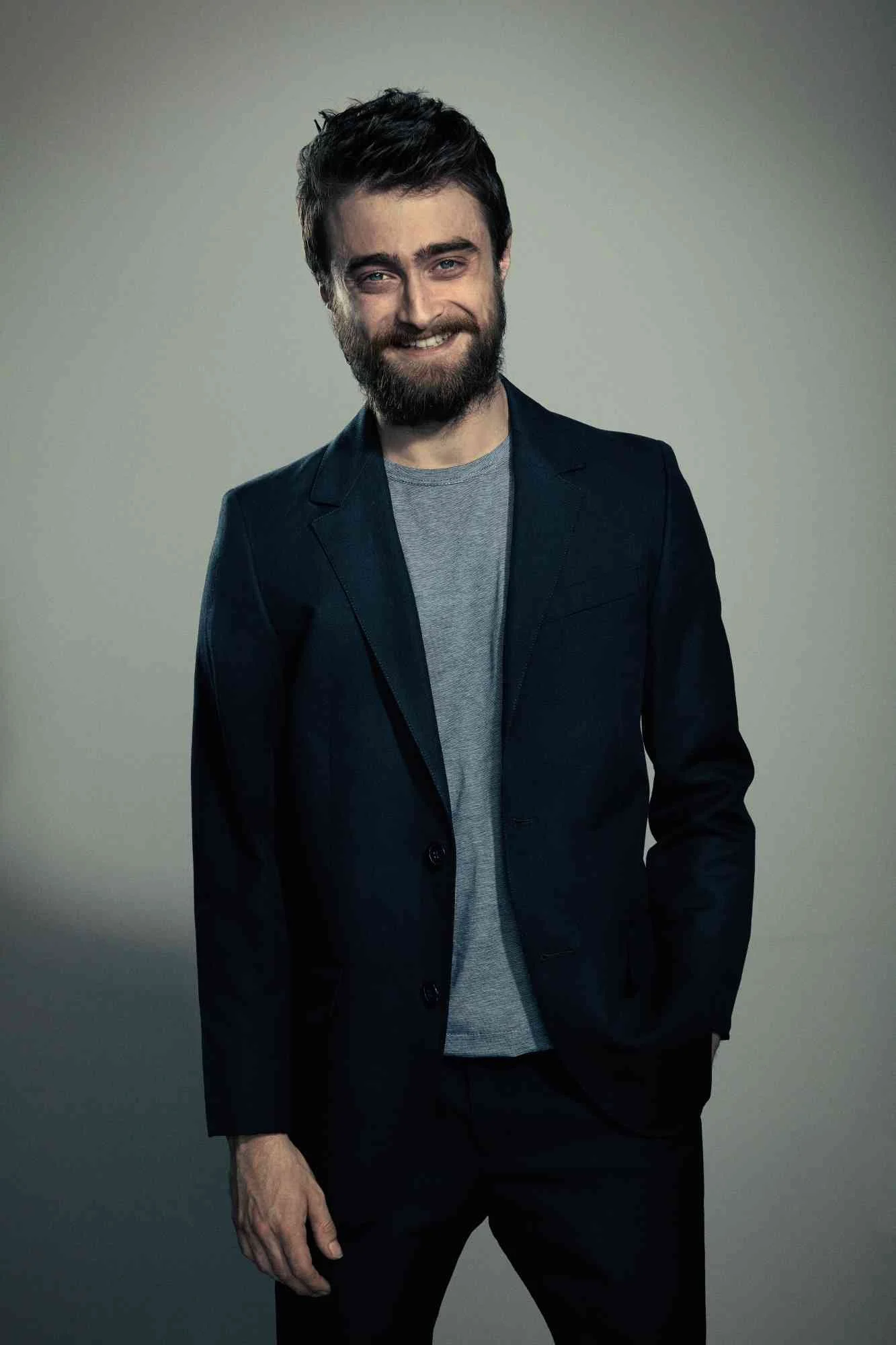 DANIEL RADCLIFFE with dark hair, beard, and blue eyes posing in front of a neutral background, wearing a dark blazer over a gray shirt, smiling slightly with one hand in his pocket.