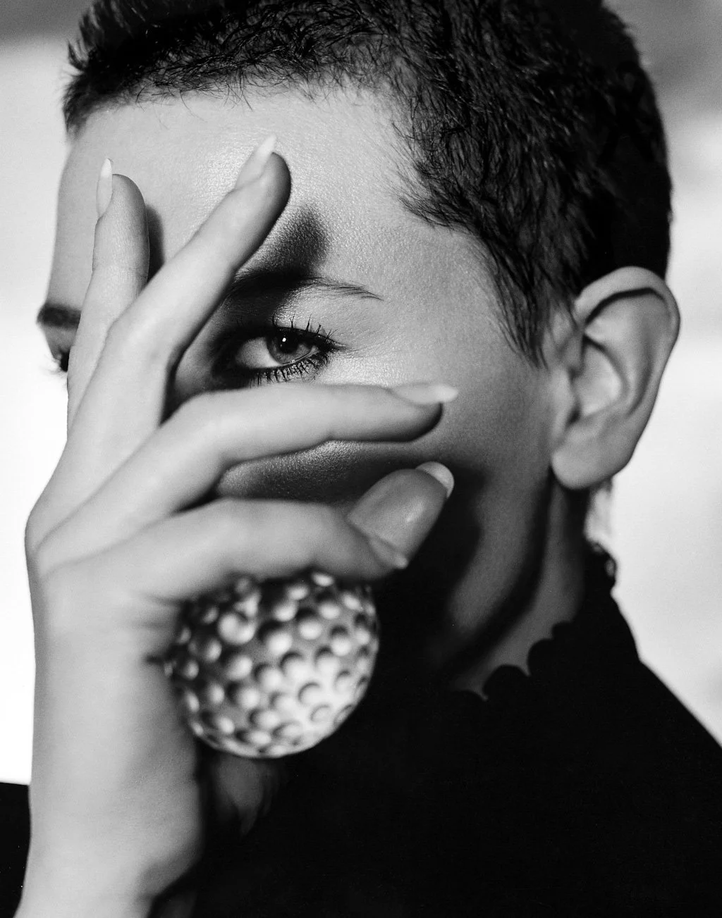 Black and white close-up of CATE BLANCHETT with short hair, covering part of their face with a hand, showcasing their eye, with a textured ring visible on their finger.