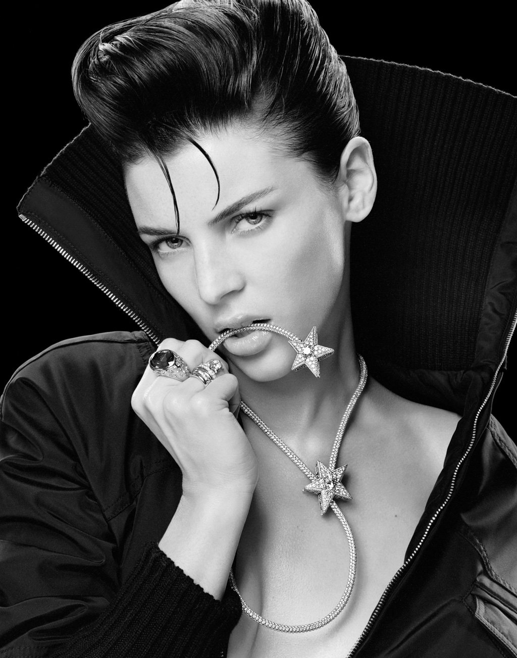 Black and white portrait of a woman with slicked-back hair, wearing a large black jacket with a high collar, holding a star-shaped necklace with a chain in her mouth, and wearing multiple rings and jewelry.