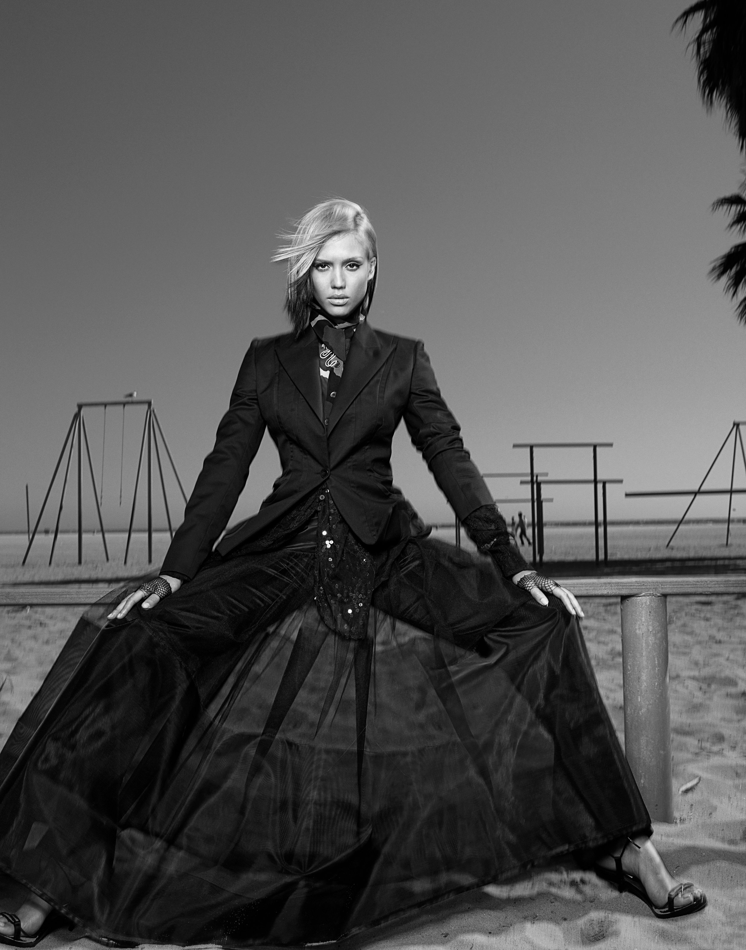 Black and white photograph of JESSICA ALBA in a black blazer and a flowing black skirt, sitting on a wooden railing on a beach with playground equipment in the background.