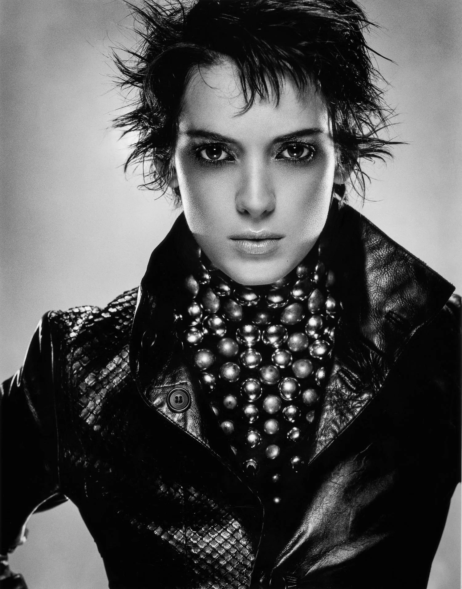 Black and white photo of WINONA RYDER with short, spiky hair, wearing a leather jacket and a large bead necklace.