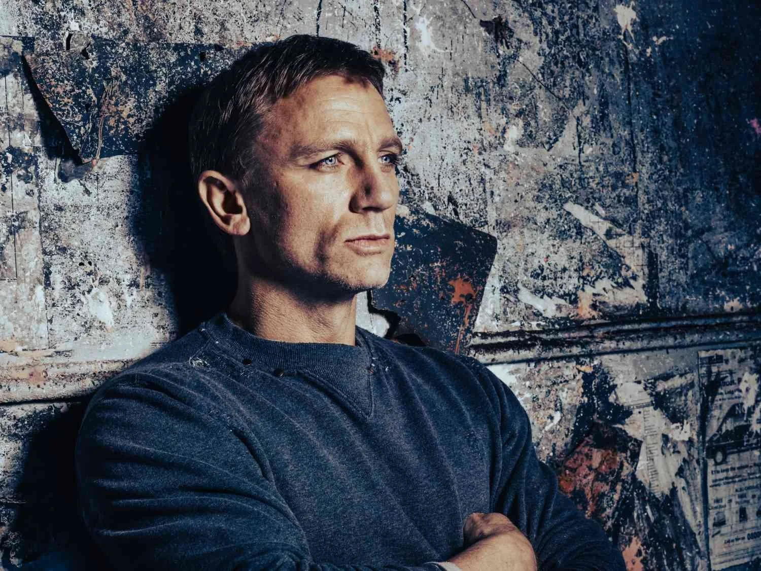 DANIEL CRAIG with short brown hair and blue eyes leaning against a graffiti-covered wall, wearing a dark sweatshirt.
