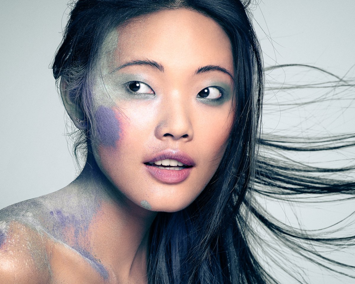 A close-up portrait of an Asian woman with long, flowing black hair, styled with colorful makeup and body paint on her shoulder and face.