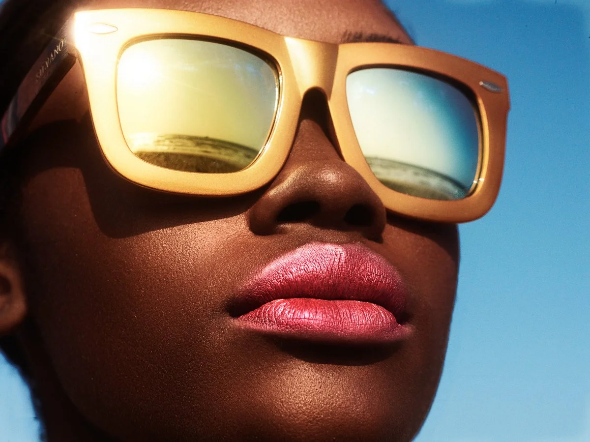 Close-up of a woman wearing beige sunglasses reflecting a beach scene, with lips slightly parted and blue sky in the background.