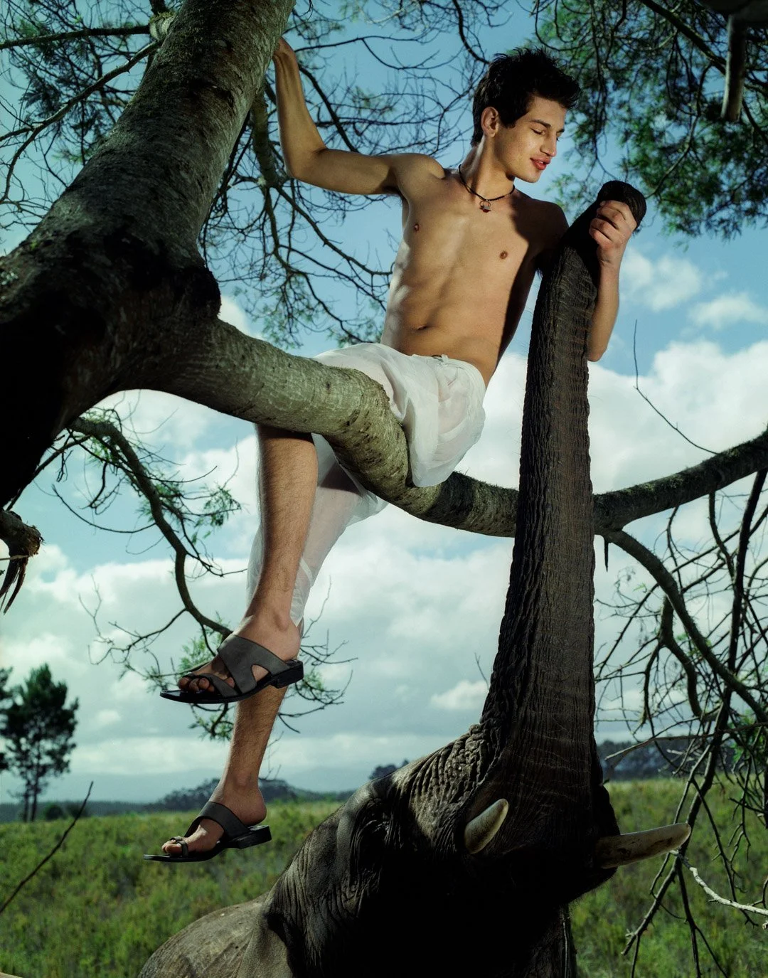 A shirtless young man with short dark hair, wearing white shorts and black sandals, sitting on a low tree branch with one arm resting on a higher branch, holding a black object, in a green and blue outdoor setting with a cloudy sky.