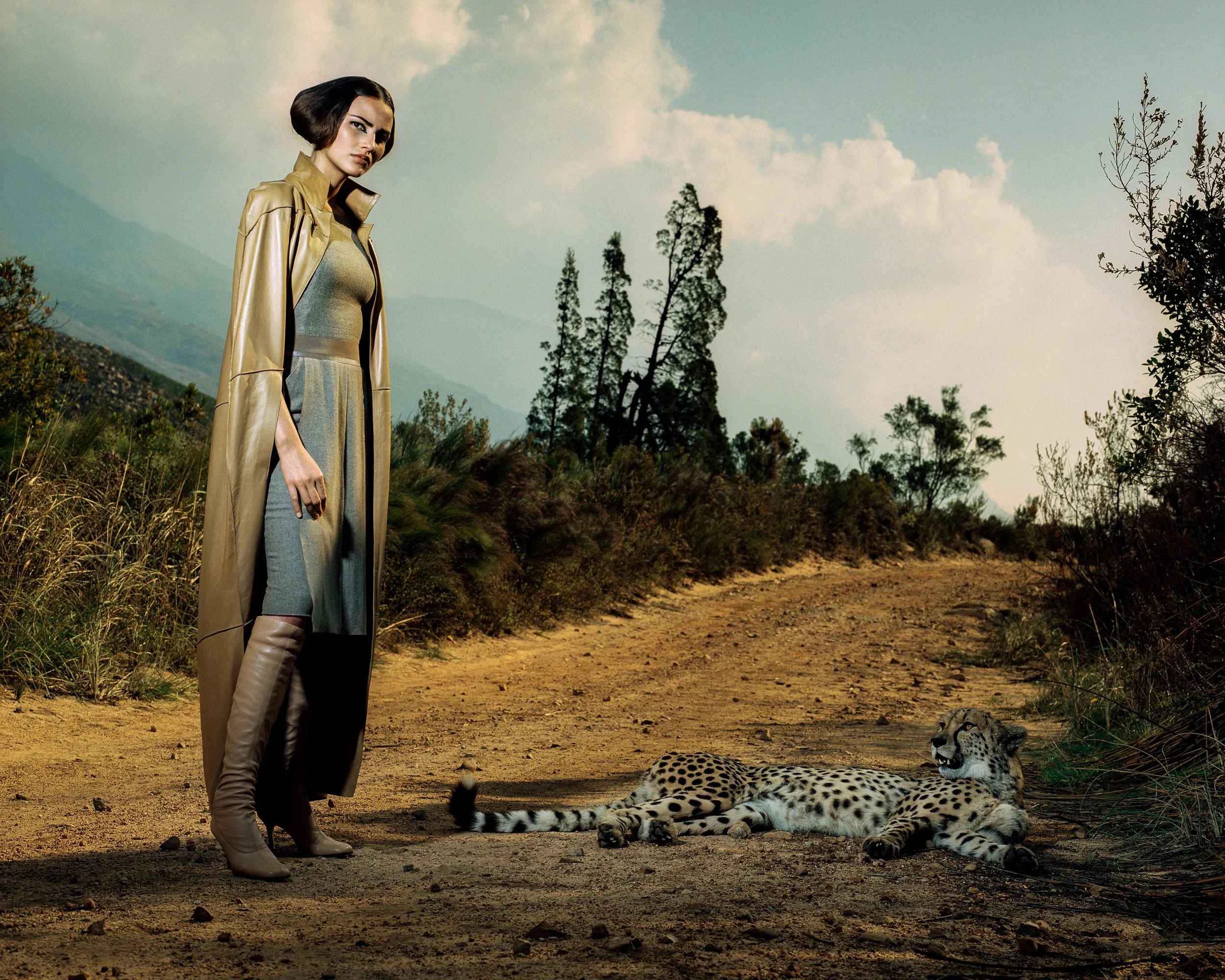 A woman dressed in a stylish trench coat and high boots standing on a dirt path in a savannah with two cheetahs lying on the ground nearby. The background features sparse bushes, tall trees, and a partly cloudy sky.