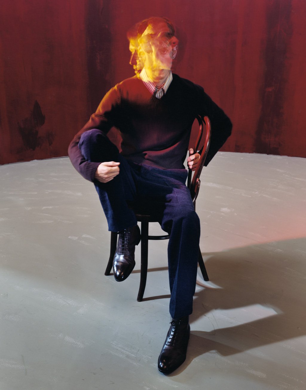 A man sitting on a chair with his right arm on his knee, wearing a dark sweater, dark pants, and shiny black shoes, with a blurred effect on his face due to a motion or double exposure, against a red background.