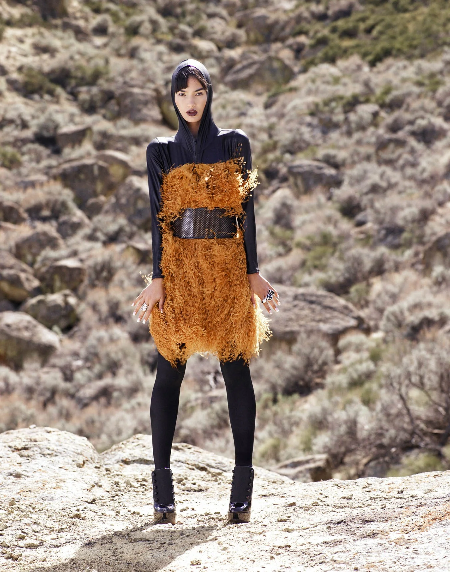 Fashion model standing outdoors in a desert landscape, wearing a black hooded top and a gold textured dress with black tights and platform shoes, accessorized with rings.