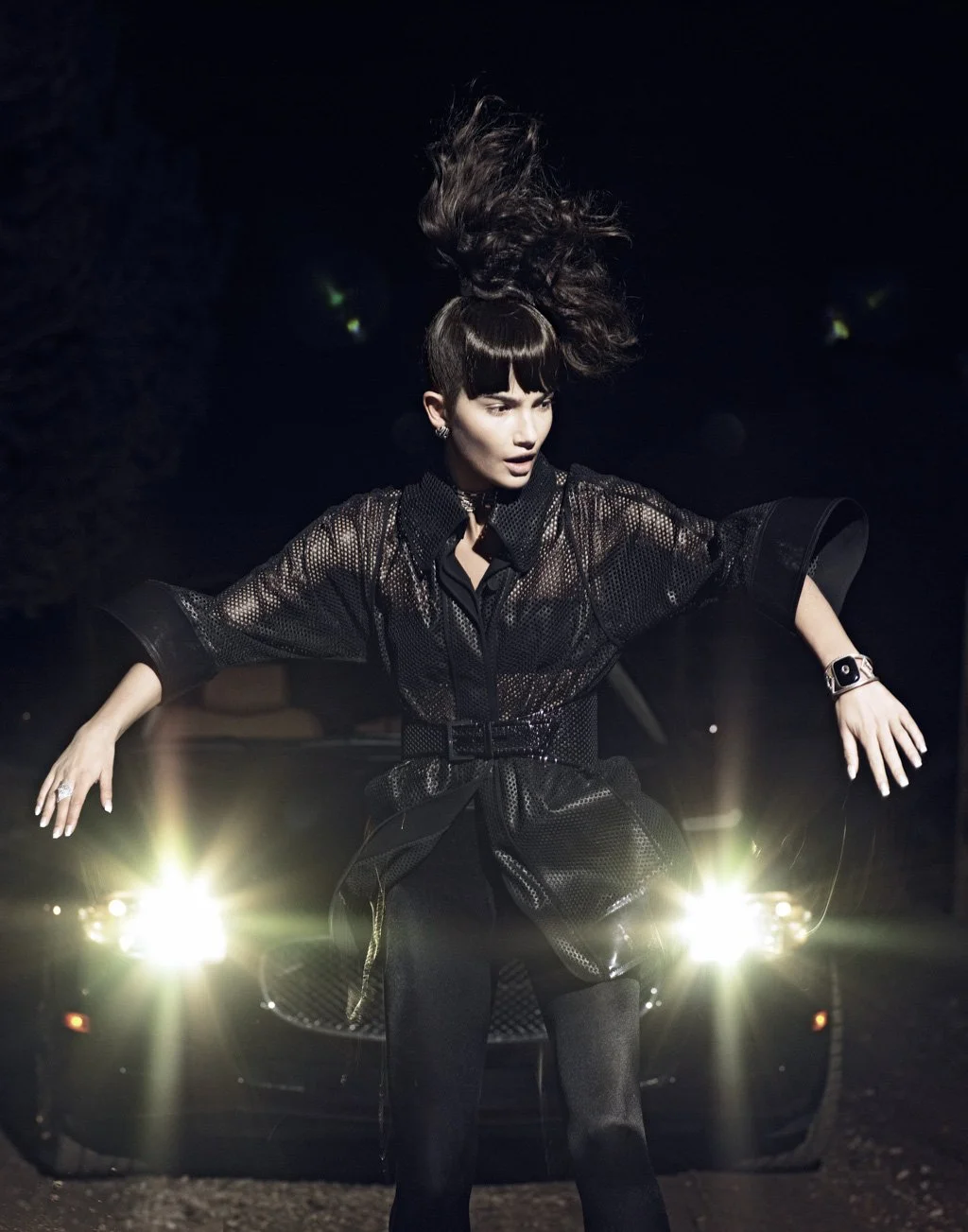 A woman standing in front of a black sports car at night, with her hair in the air and headlights shining behind her.