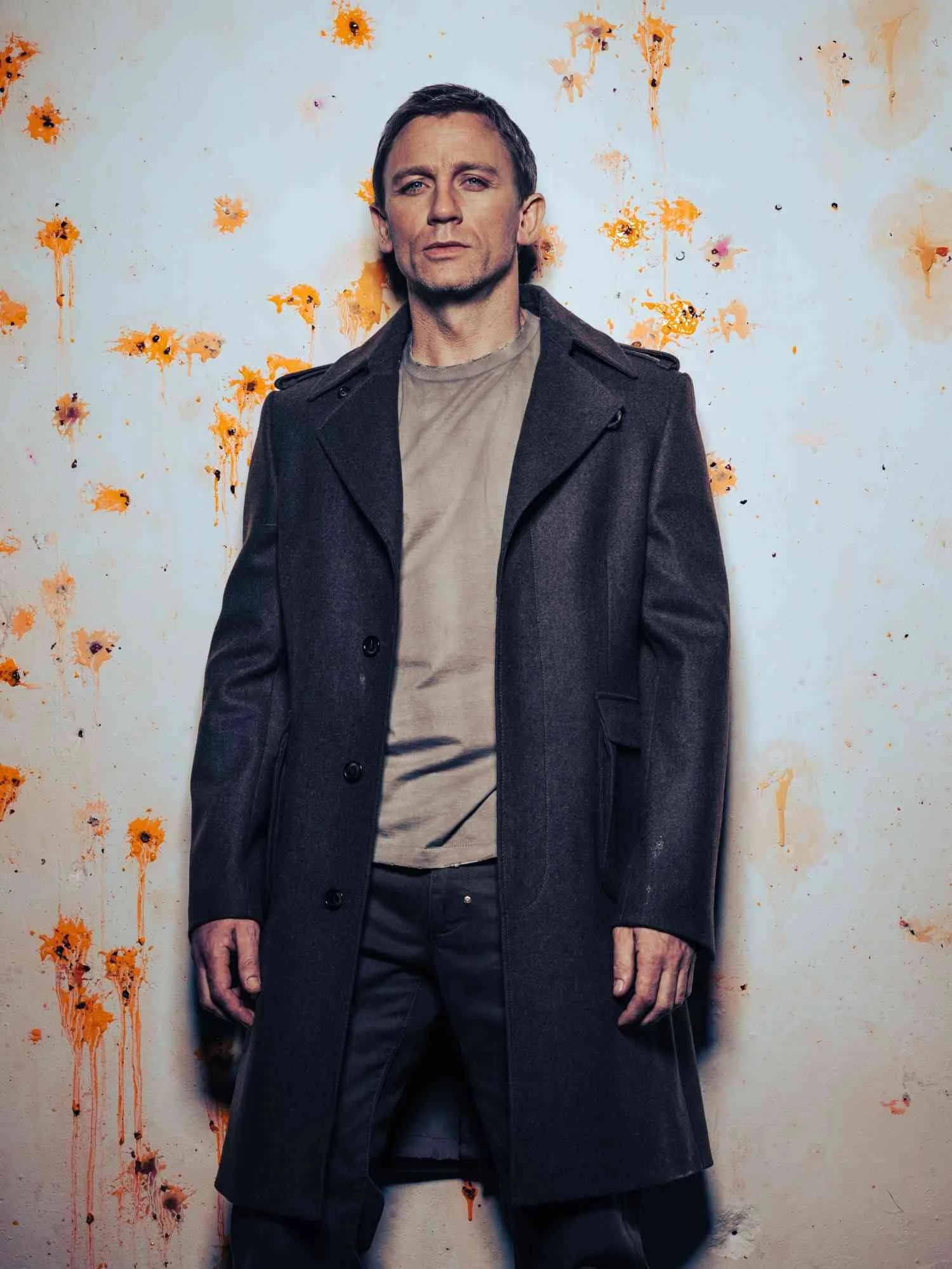 DANIEL CRAIG stands in front of a wall splattered with orange and black paint, wearing a dark coat and beige shirt.