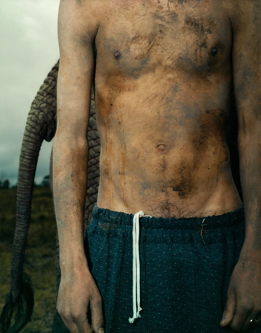 Close-up of a shirtless man's dirty, muddy torso and arms, wearing dark drawstring shorts with a background of a cloudy sky and grass field.