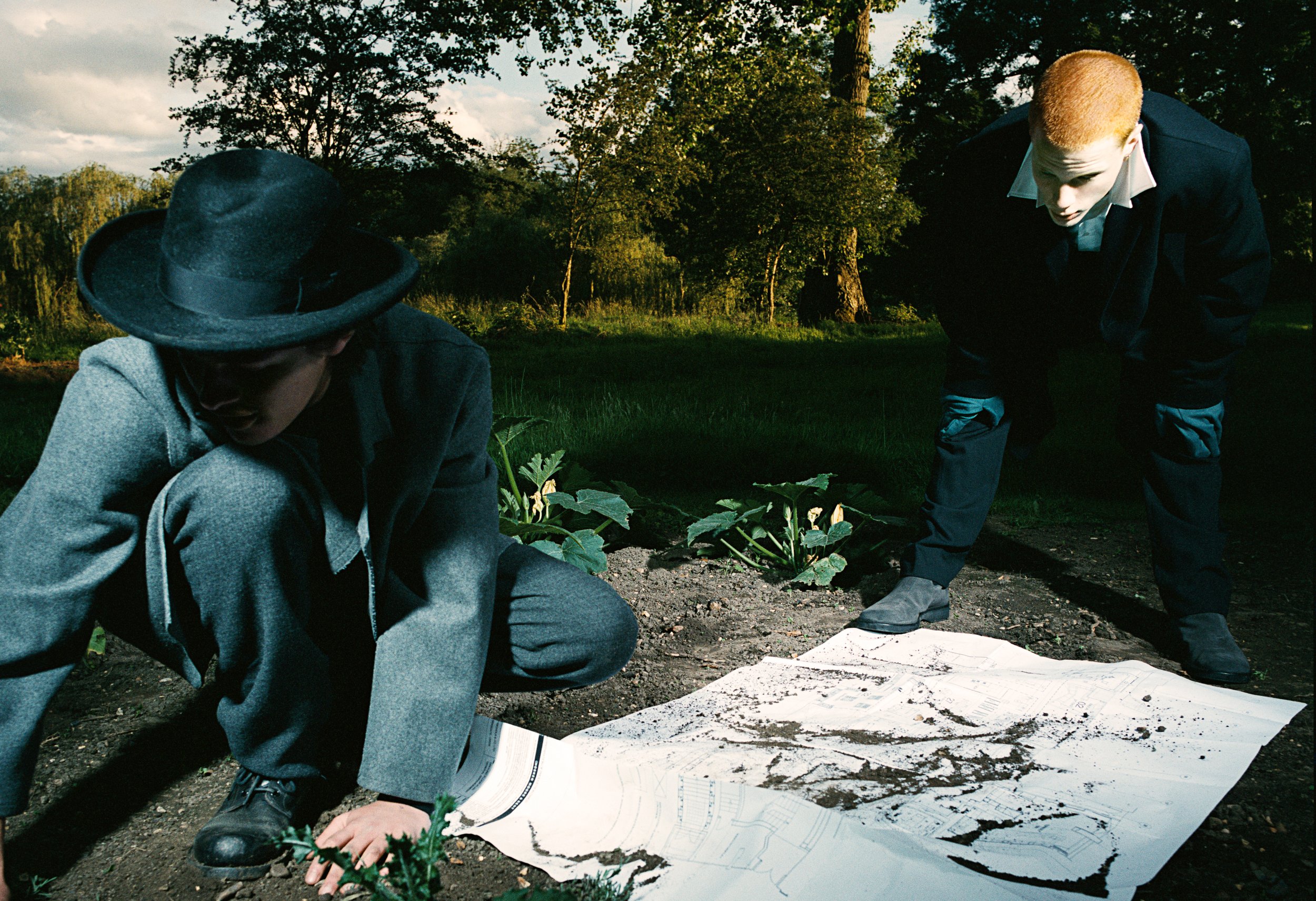 Two men in formal attire examining a site plan on the ground outdoors, with plants and trees in the background.
