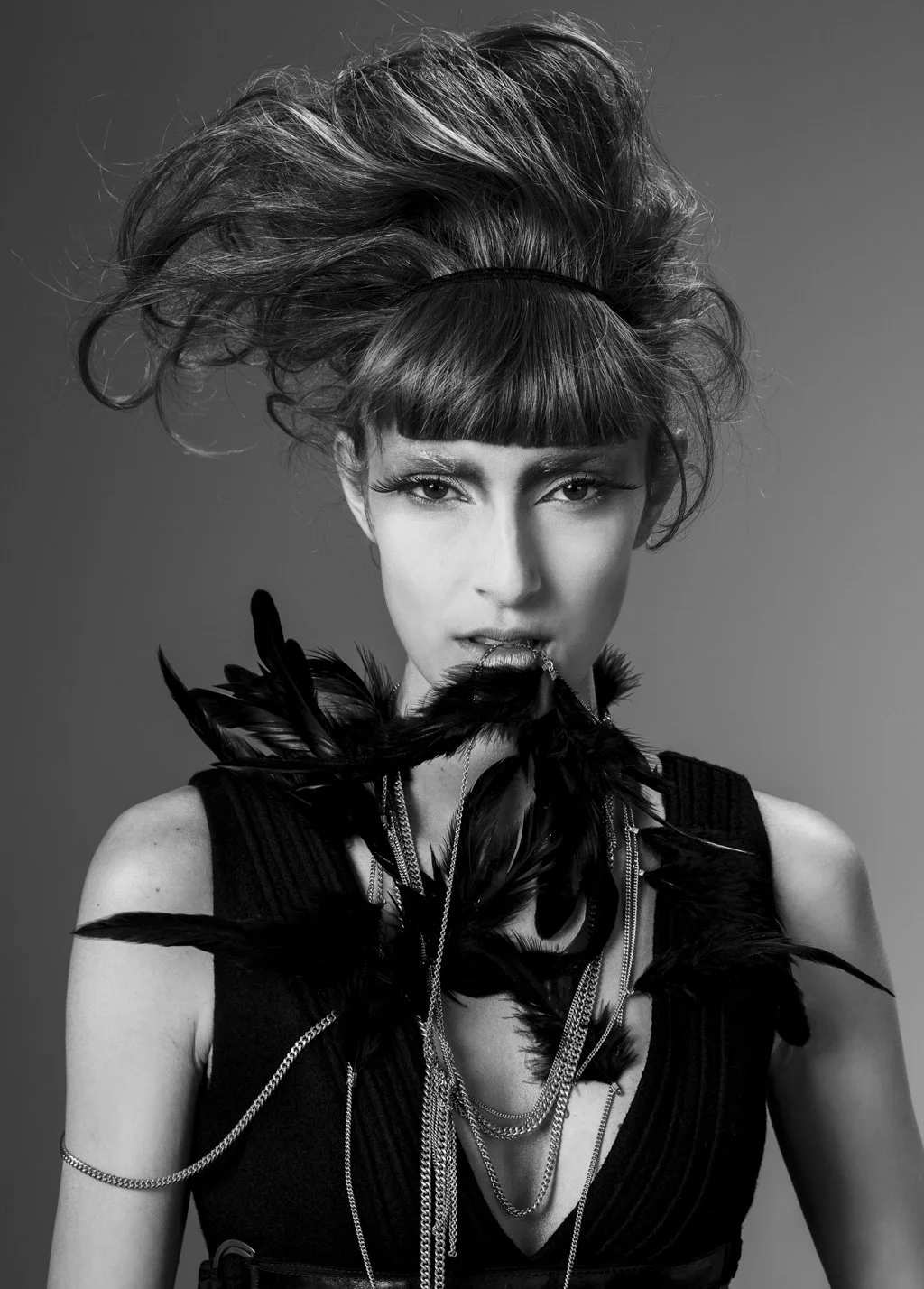 A black-and-white portrait of a woman with avant-garde hairstyle, holding black feathers in her mouth, wearing layered necklaces and a sleeveless top.