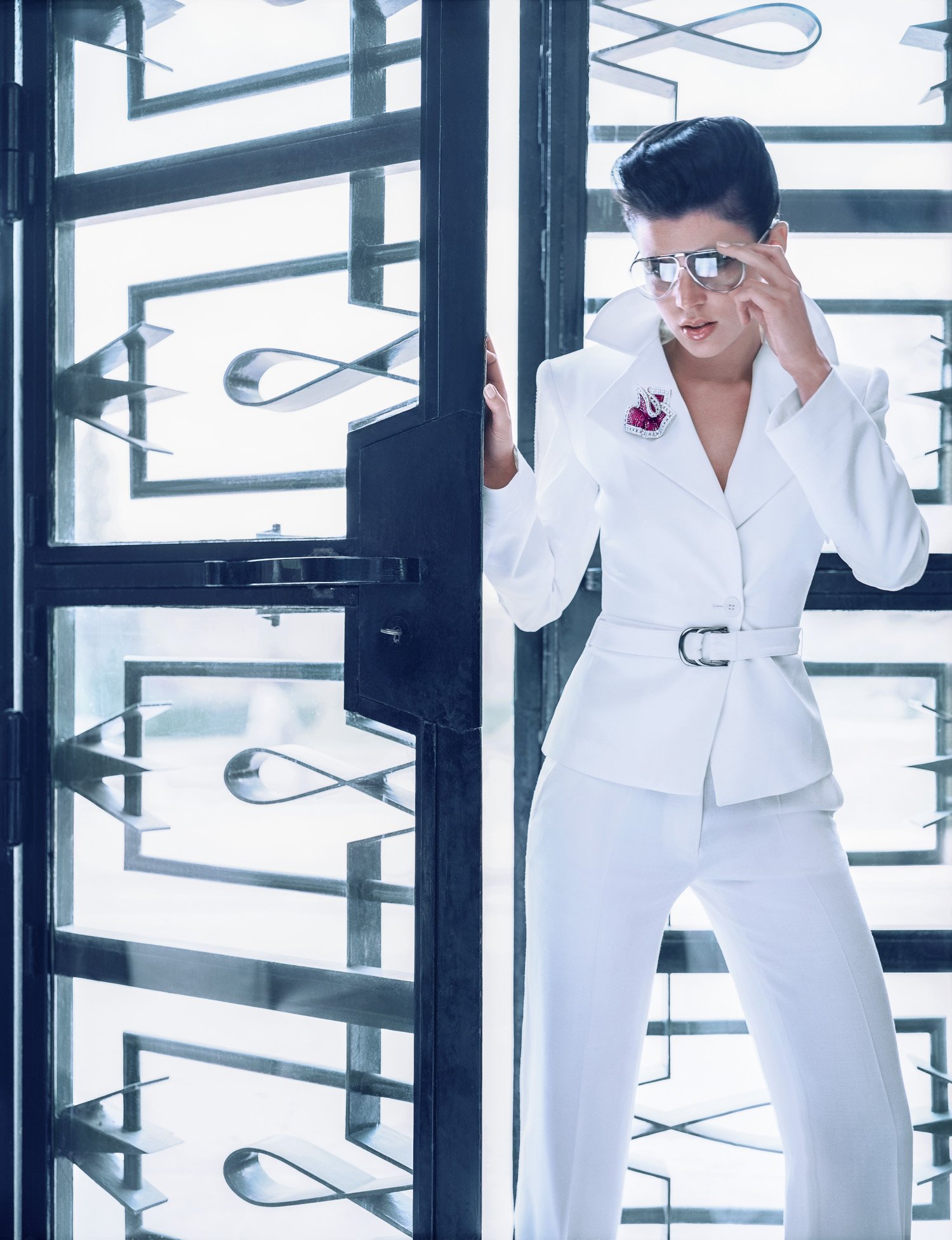 Woman in a white suit with glasses looking surprised or confused, standing near glass doors with modern metallic designs.