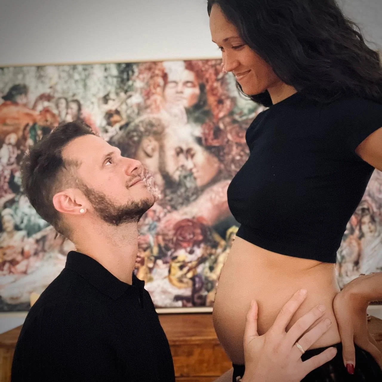 A man kneeling and looking up at a woman who is sitting on a surface, holding her waist and smiling at him with a colorful painting in the background.