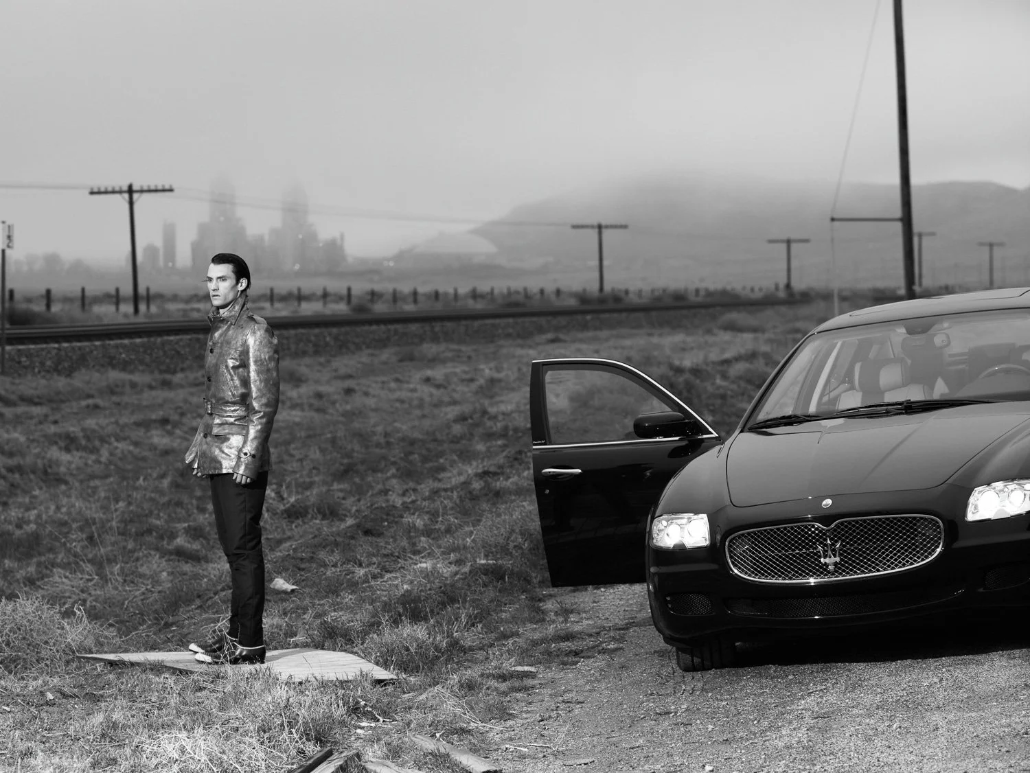 A man in a leather jacket standing on a grassy roadside next to a black Maserati with the door open, in a rural area with train tracks and power lines, with a city skyline and misty hills in the background.