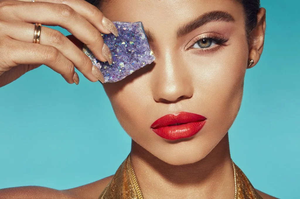A woman with bright red lipstick and gold earrings holds a decorative purple and silver makeup sponge to her eye against a turquoise background.