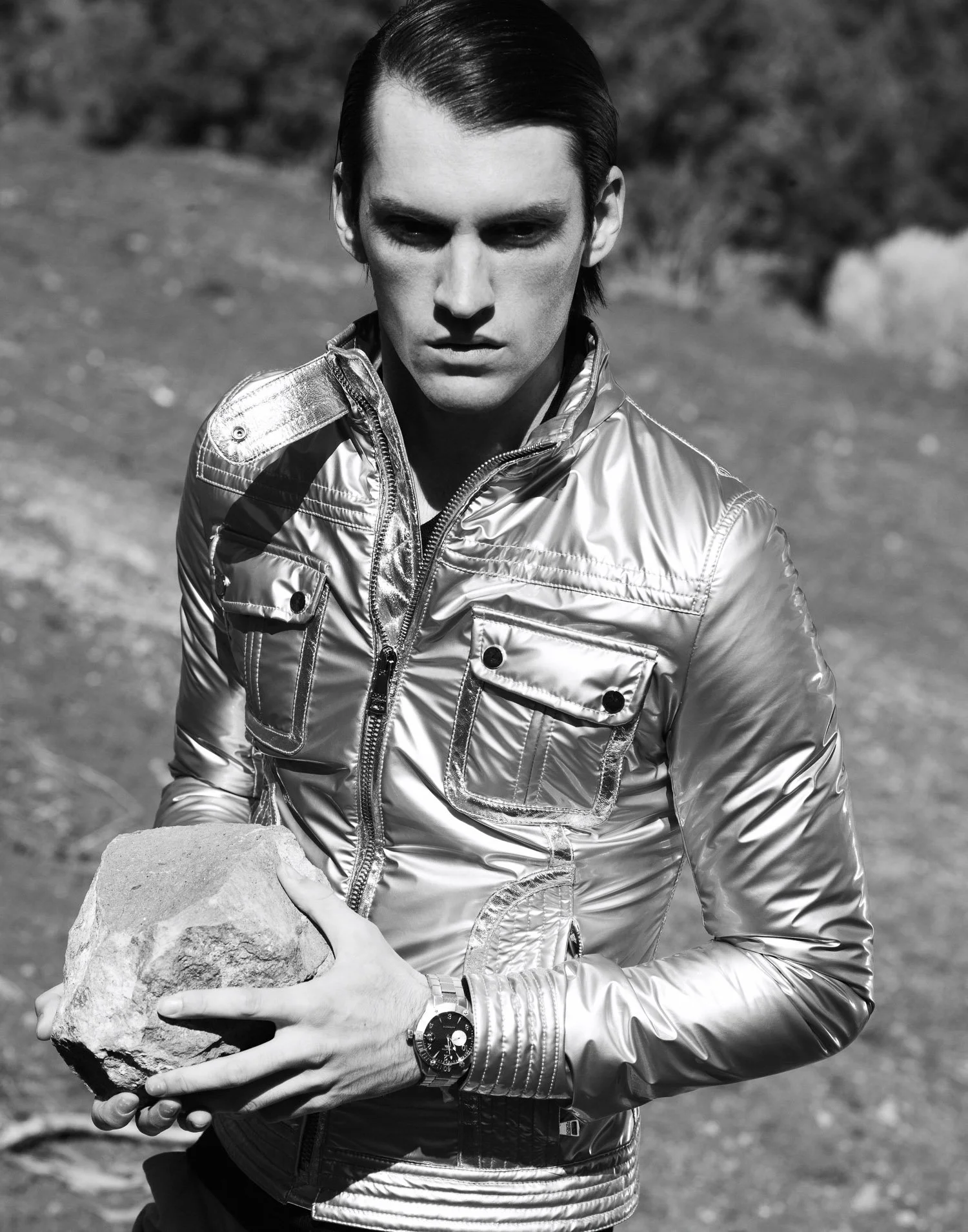 Black and white photo of a man wearing a shiny, metallic jacket, holding a large rock in his hands outdoors.