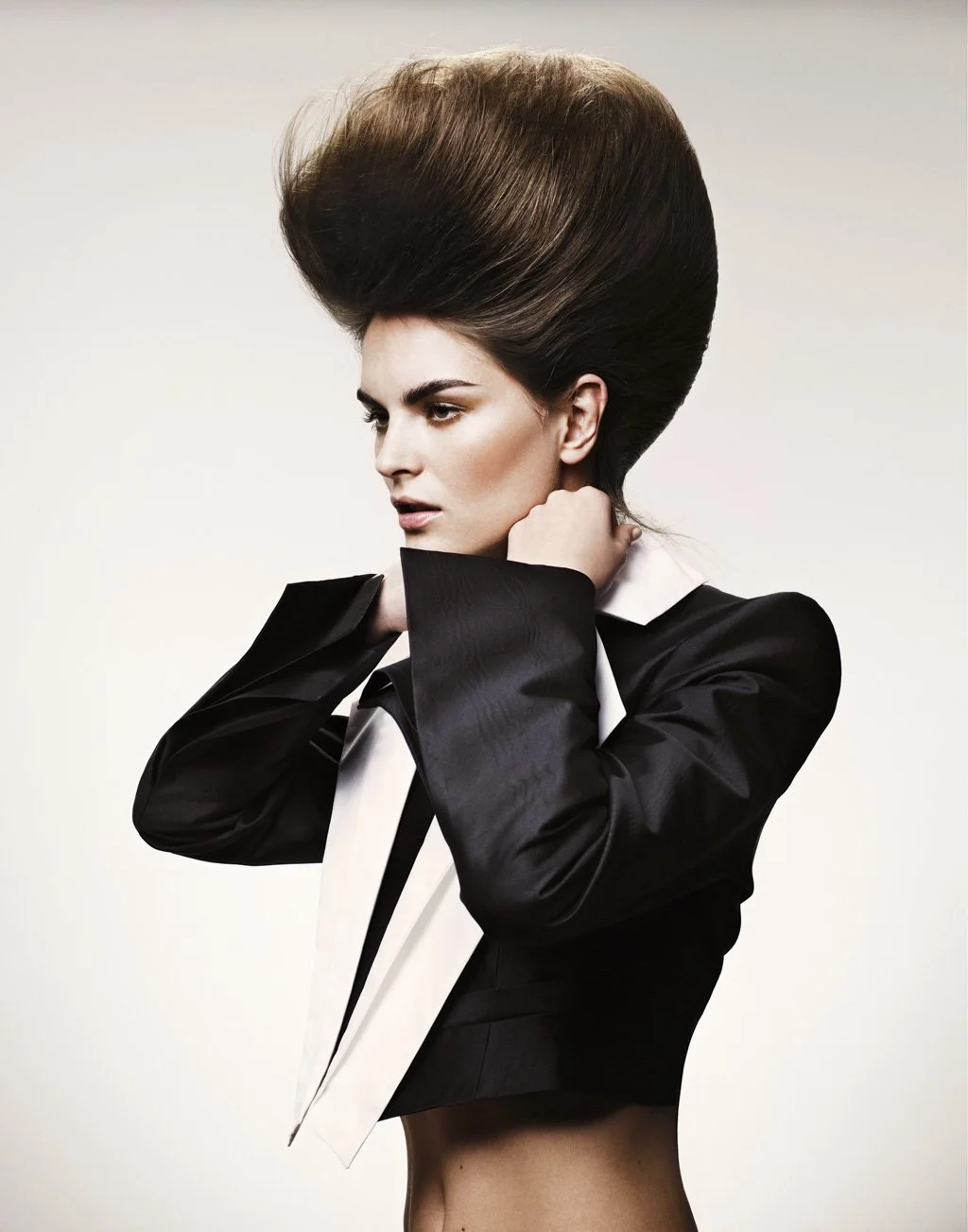 A woman with tall, voluminous hair styled in a retro fashion, wearing a black blazer with rolled-up sleeves and a white shirt, posing against a plain background.